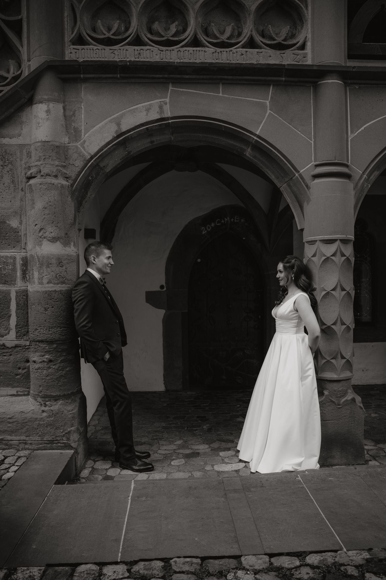 Black and white photo of bride and groom standing apart smiling under a gothic stone archway.