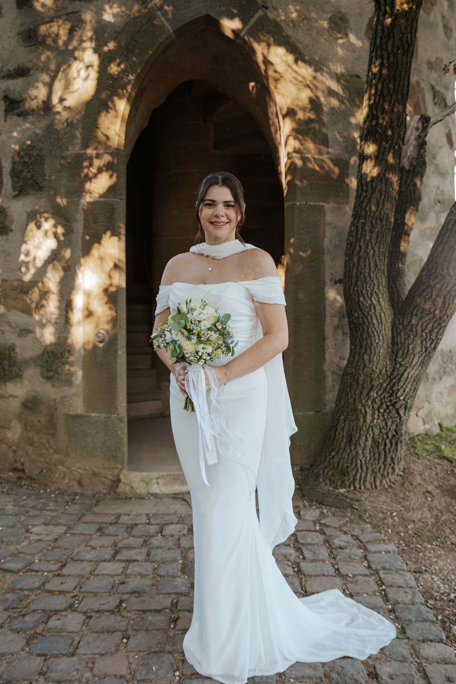 Beautiful bride in fitted off-shoulder white gown holding bouquet while standing in stone arch doorway.