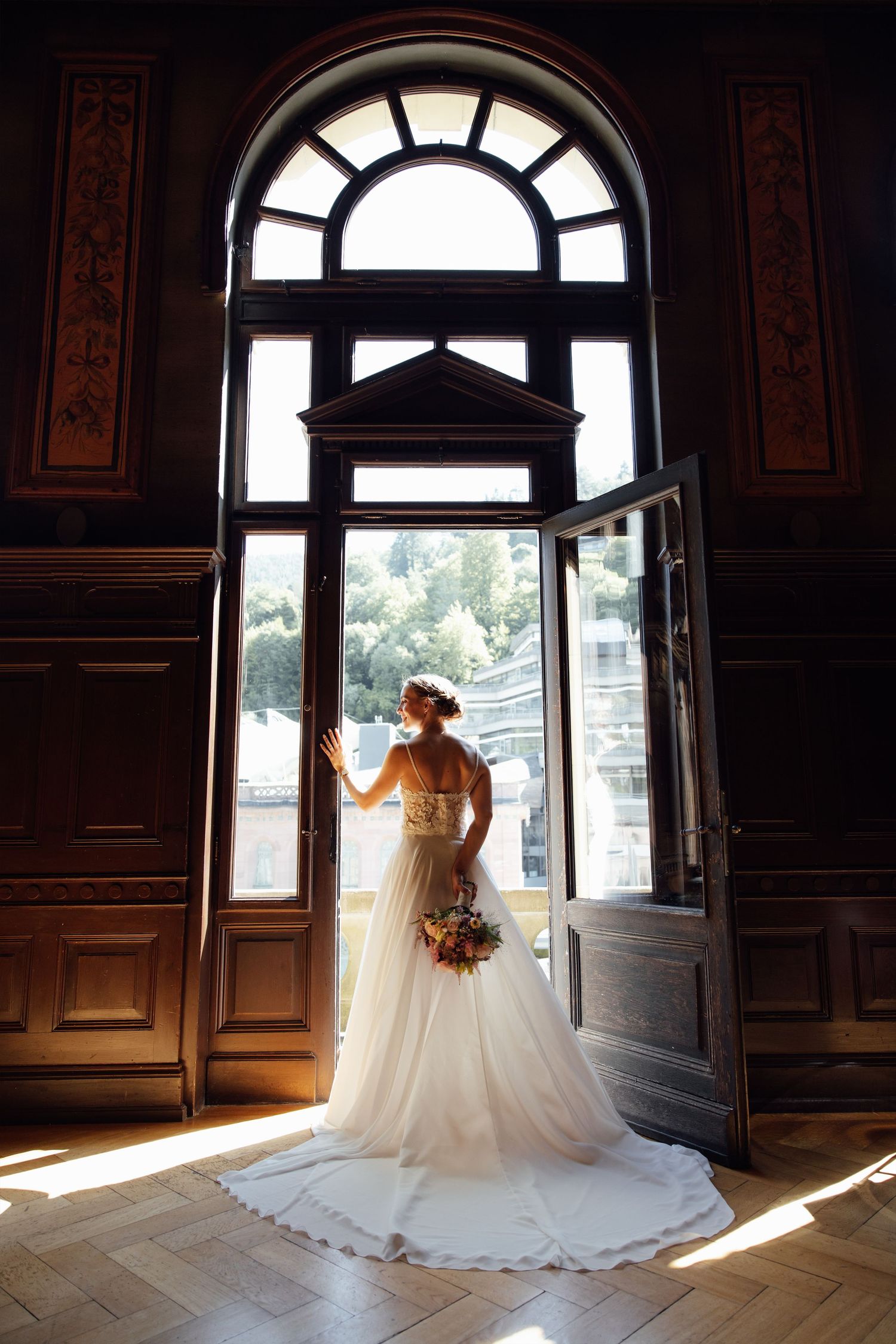 Bride in white gown stands at ornate wooden doors, holding bouquet, bathed in sunlight from arched window.