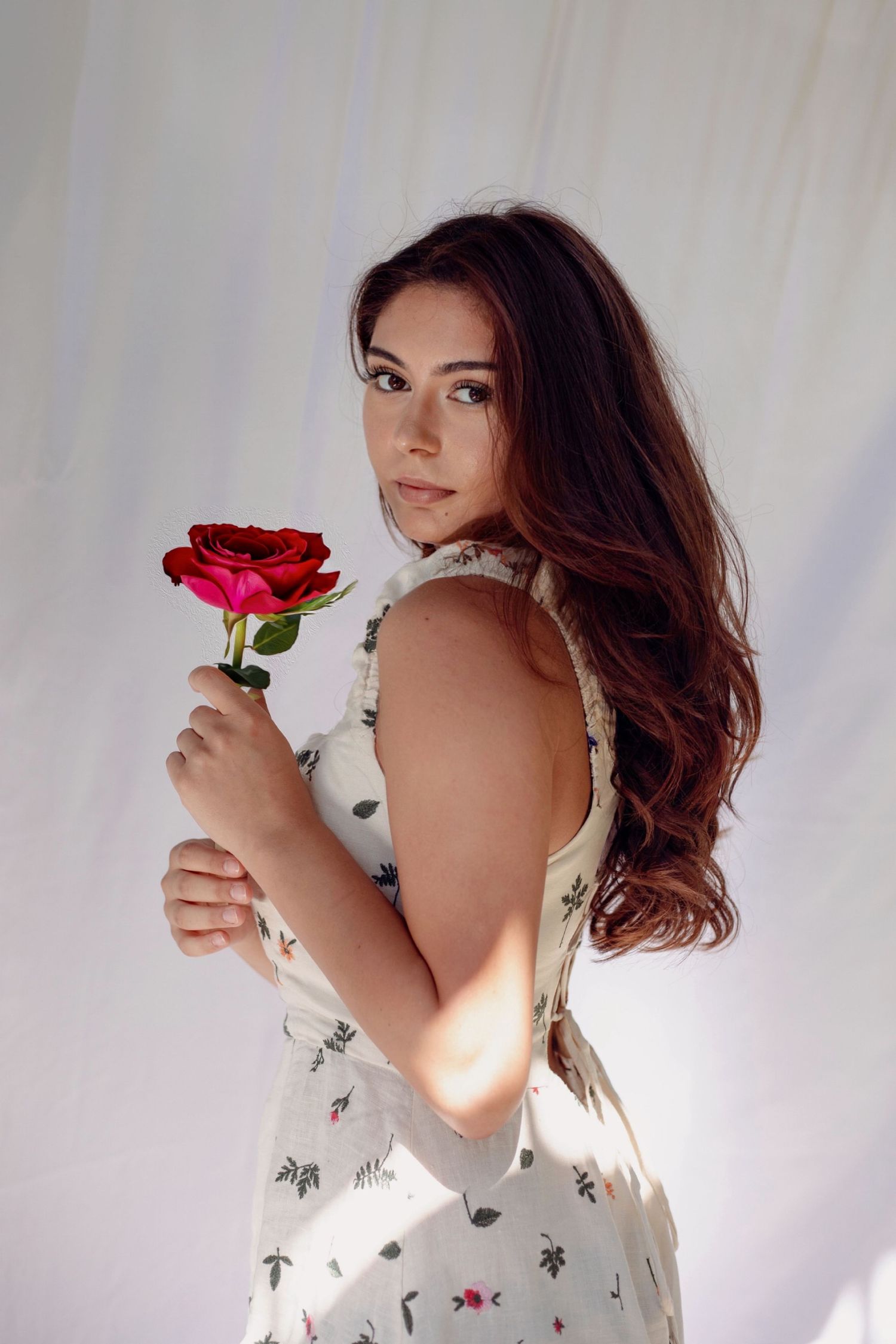 Brunette woman in a floral dress holding a red rose while looking over her shoulder indoors.