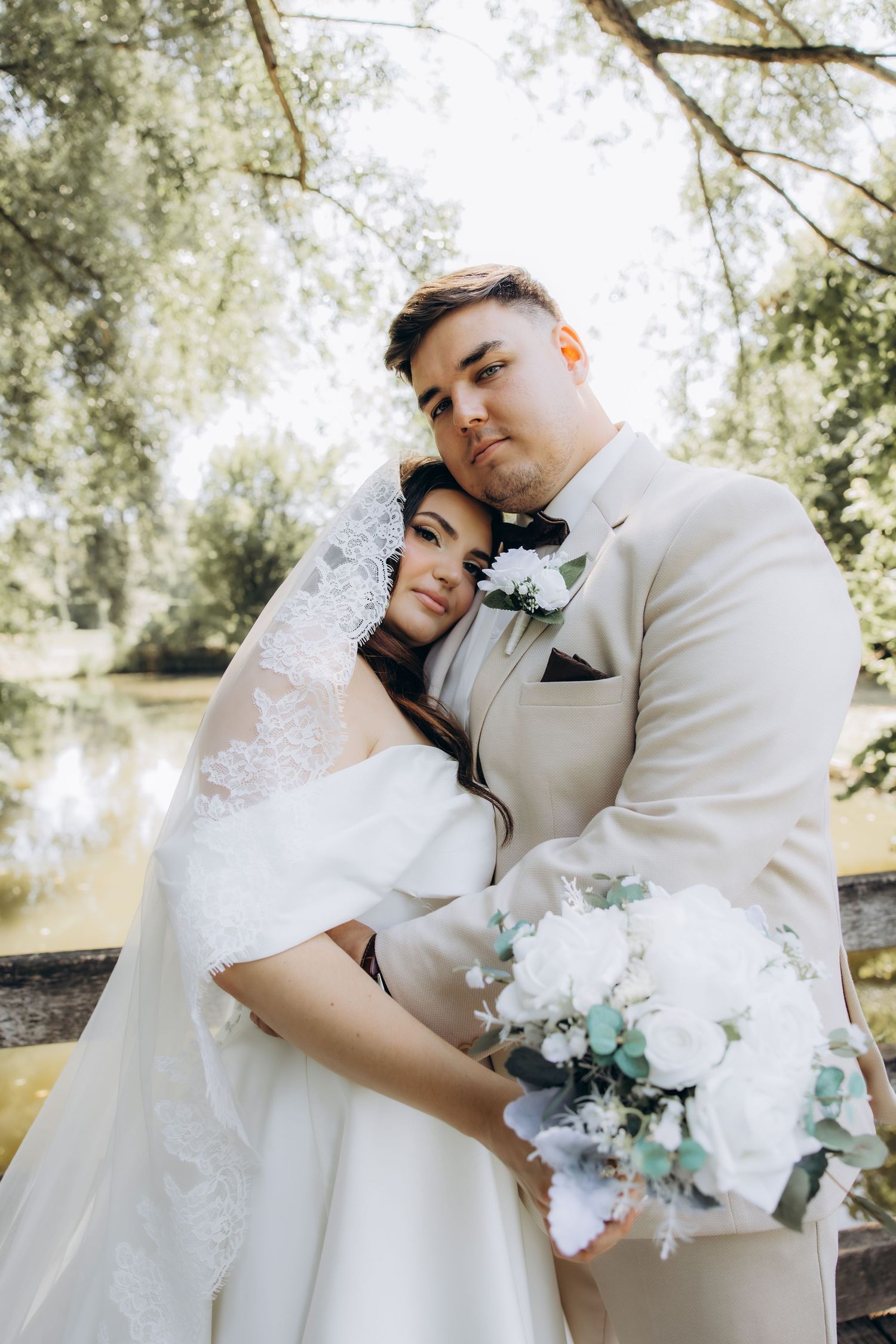 A bride in a white lace gown and veil embraces her groom in a beige suit outdoors, holding a white floral bouquet.