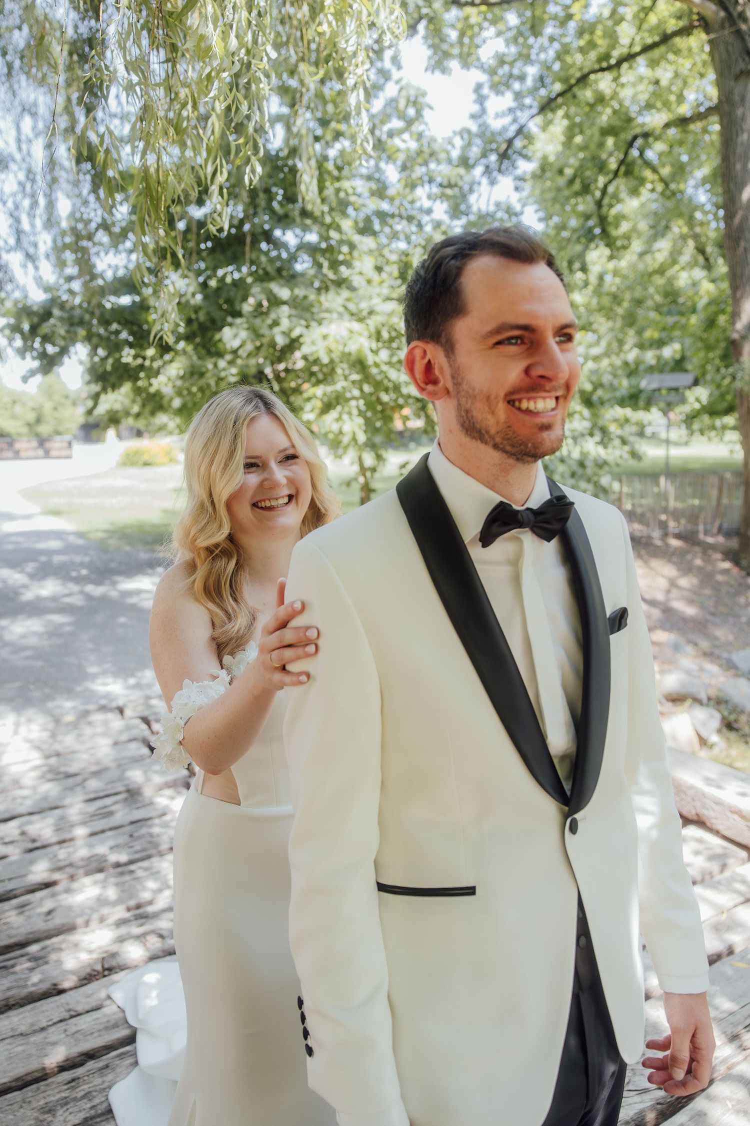 Smiling bride in white strapless gown touches grooms shoulder as he laughs in cream tuxedo outdoors.