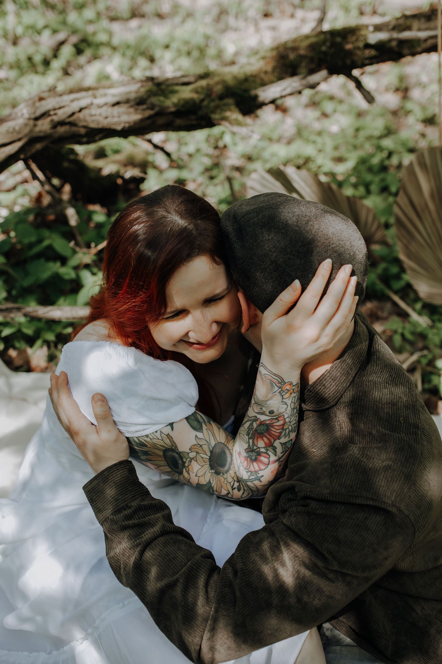 Couple embracing in forest, bride in white dress with tattooed arms hugging groom in dark jacket.