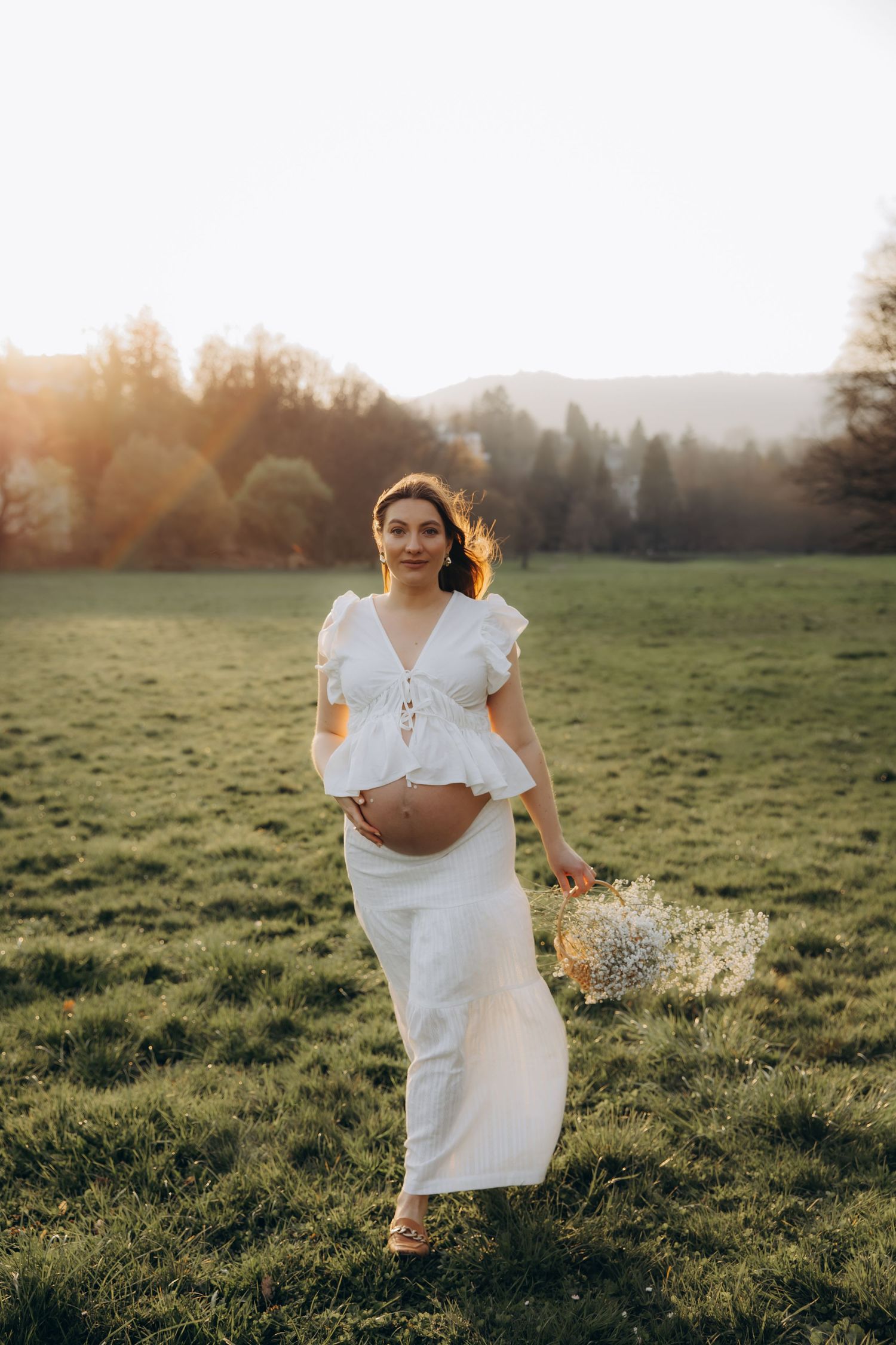 Smiling pregnant woman in white dress holding wildflowers while standing in a sunlit mountain meadow.