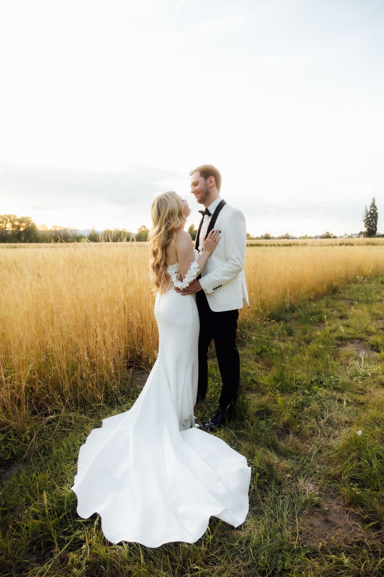 Bride and groom embrace in golden field at sunset, bride in elegant white gown with long train.