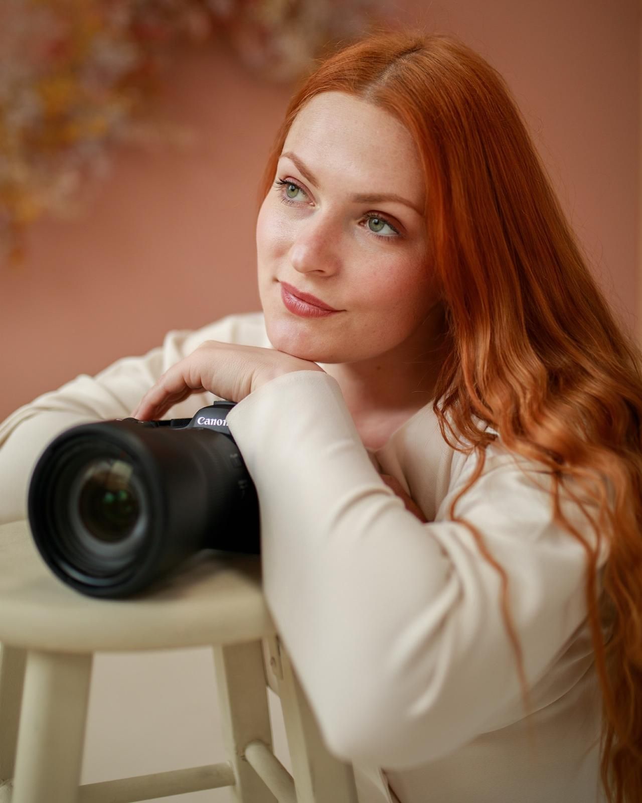 A red-haired woman with green eyes poses with a camera on a stool in a soft pink studio setting.