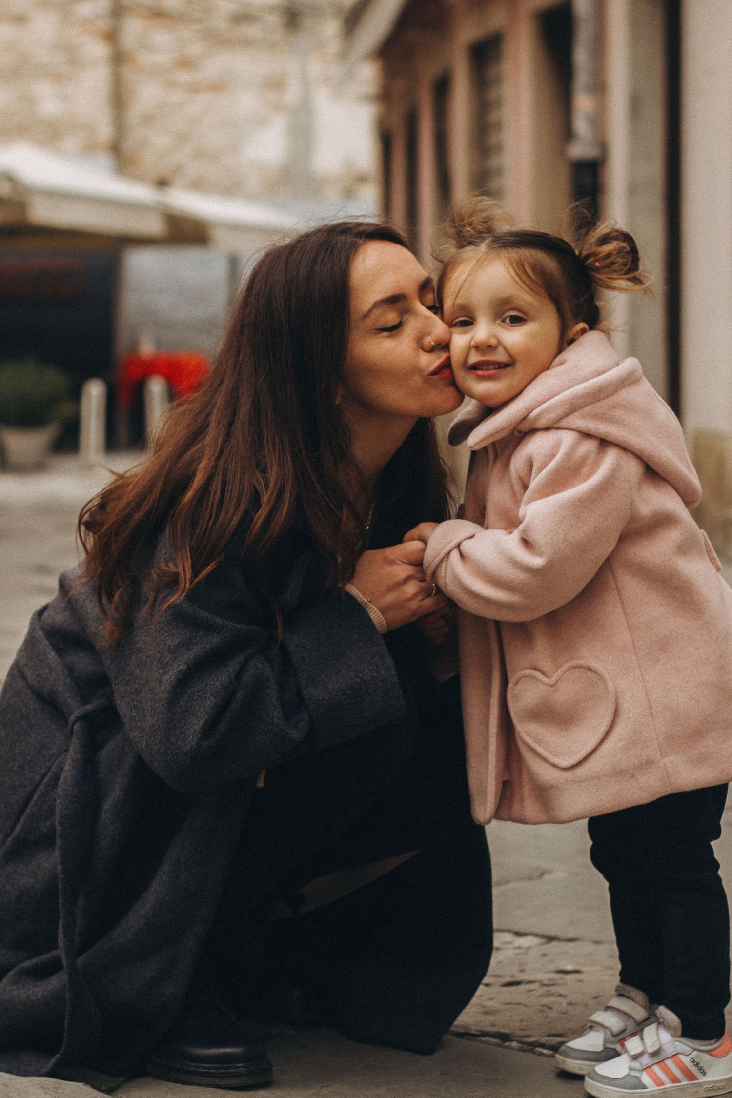 Mother crouching to kiss her smiling toddler daughter on a city street.