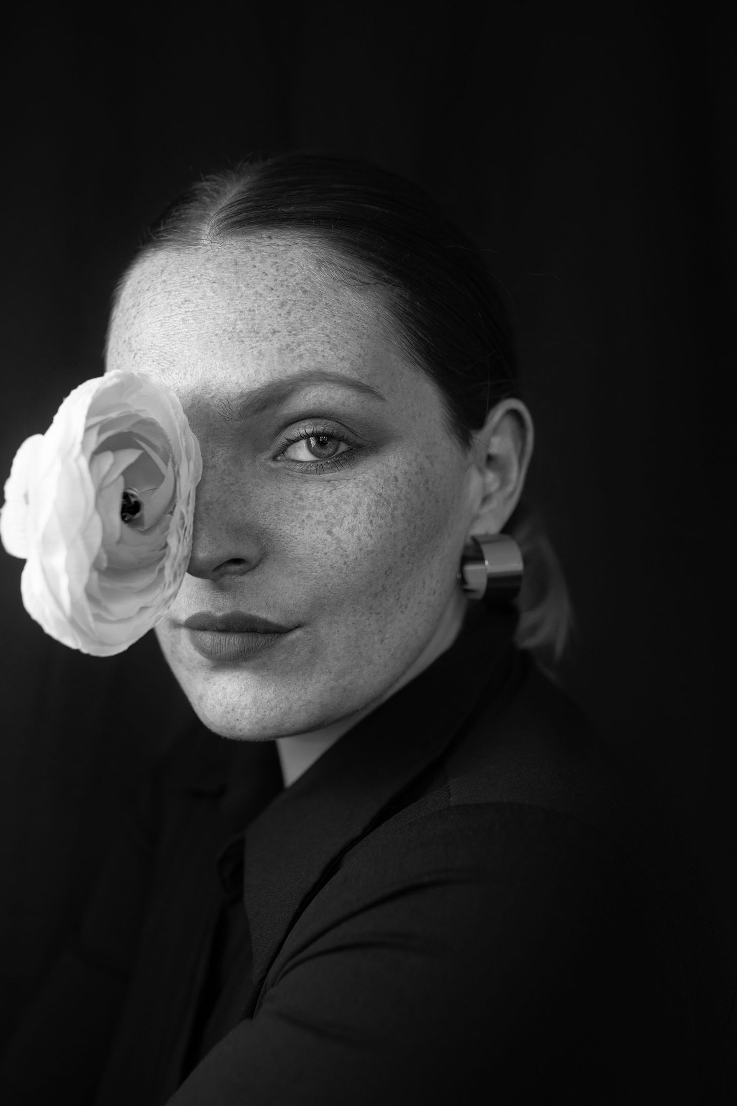 Black and white portrait of a freckled woman holding a white ranunculus flower over one eye.