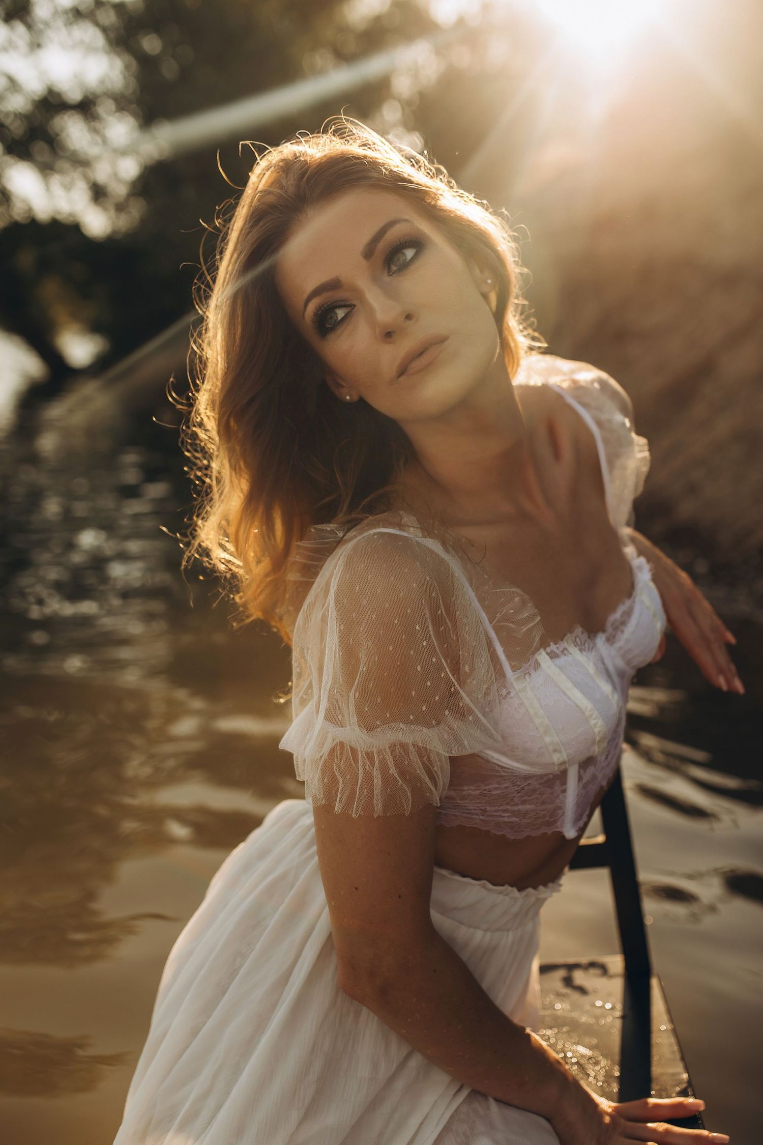 A woman in a white vintage dress posing near water in warm golden backlit sunlight.