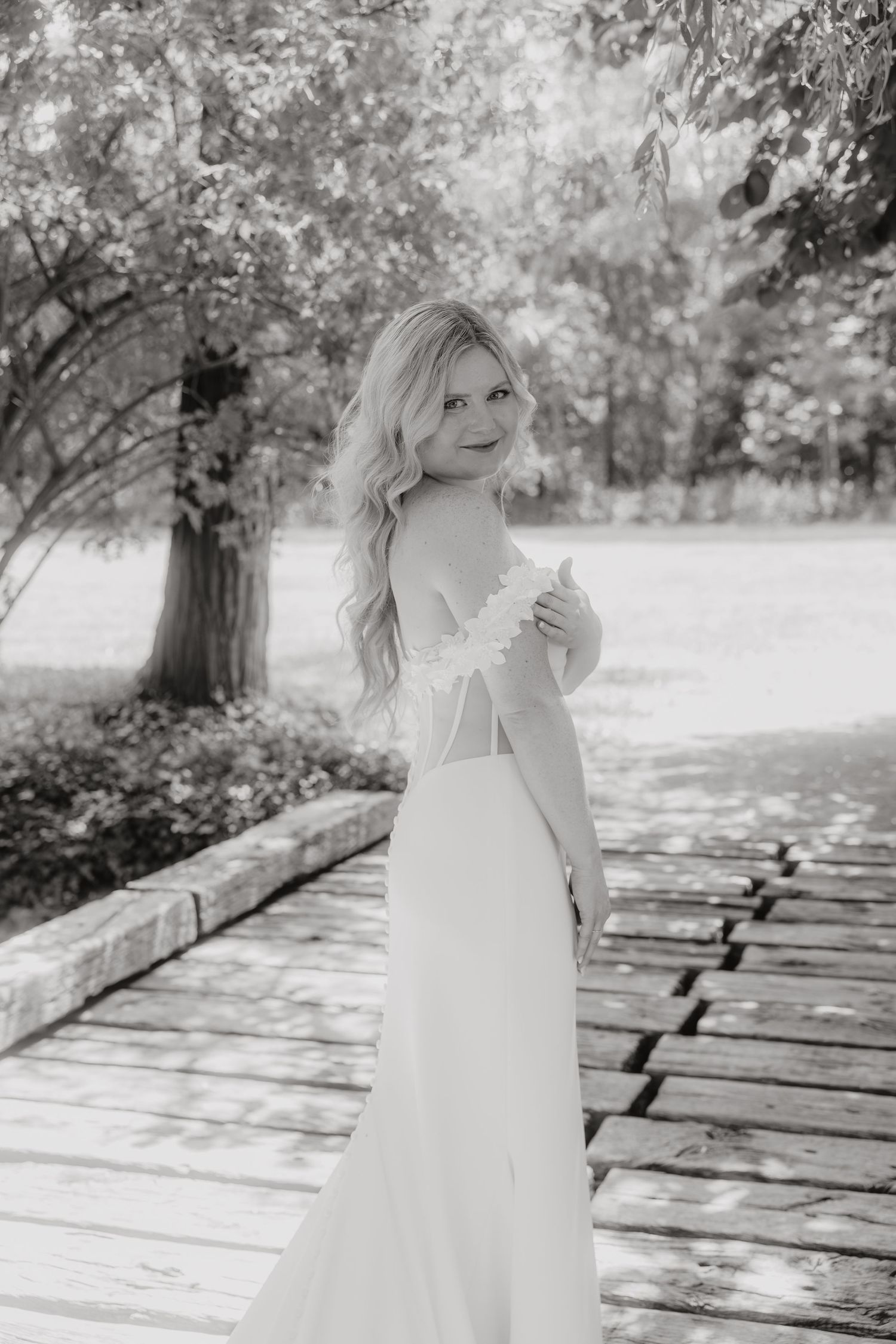 Black and white photo of a blonde bride in an elegant backless wedding gown posing on a wooden path surrounded by trees.