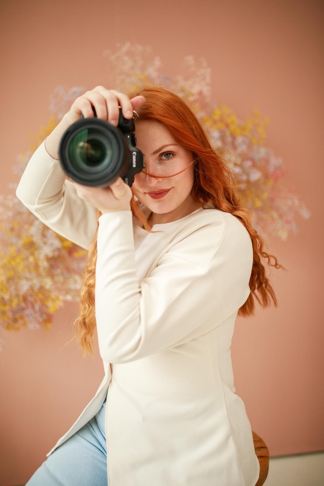 Smiling redhead female photographer holding a Canon camera up to her eye in a pink floral studio.