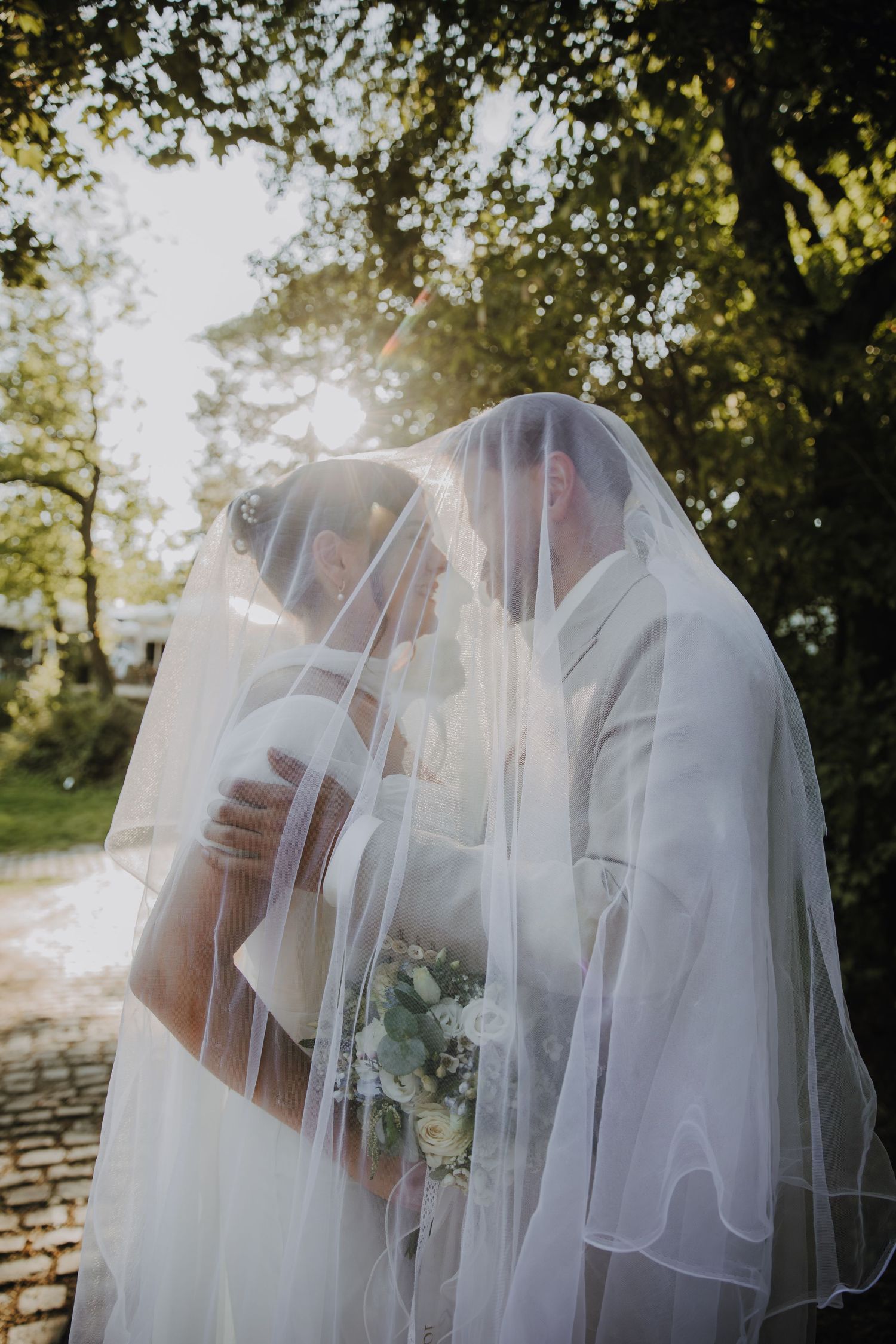 Bride and groom sharing romantic kiss under flowing white veil in golden sunlight among green trees.