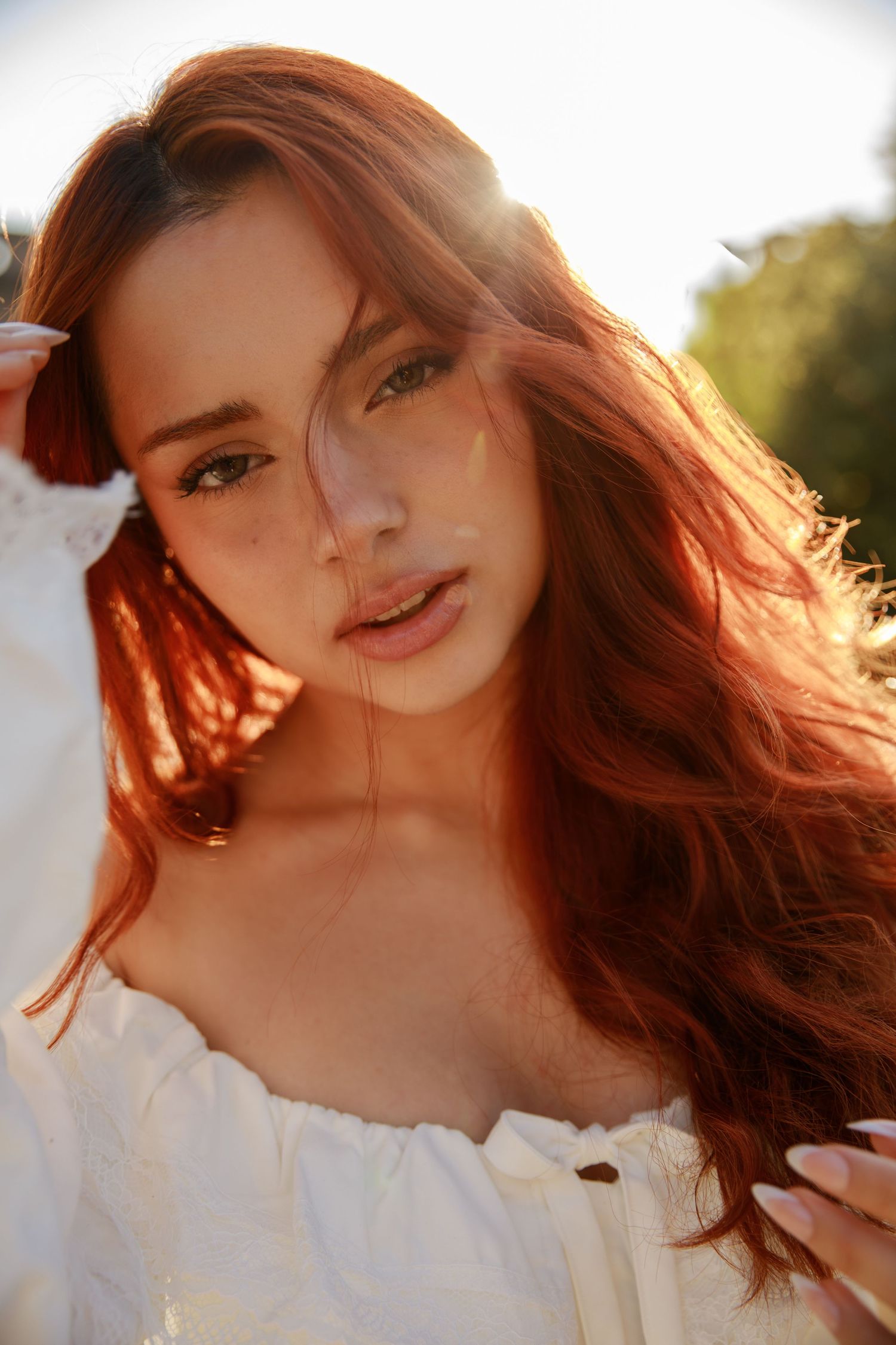 Young woman with long red hair wearing white off-shoulder top poses outdoors in golden sunlight.