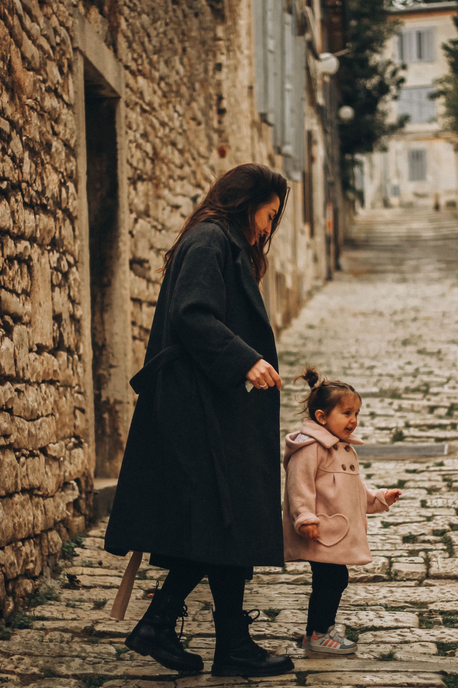 Mother and young daughter walking together on a cobblestone old town alley.