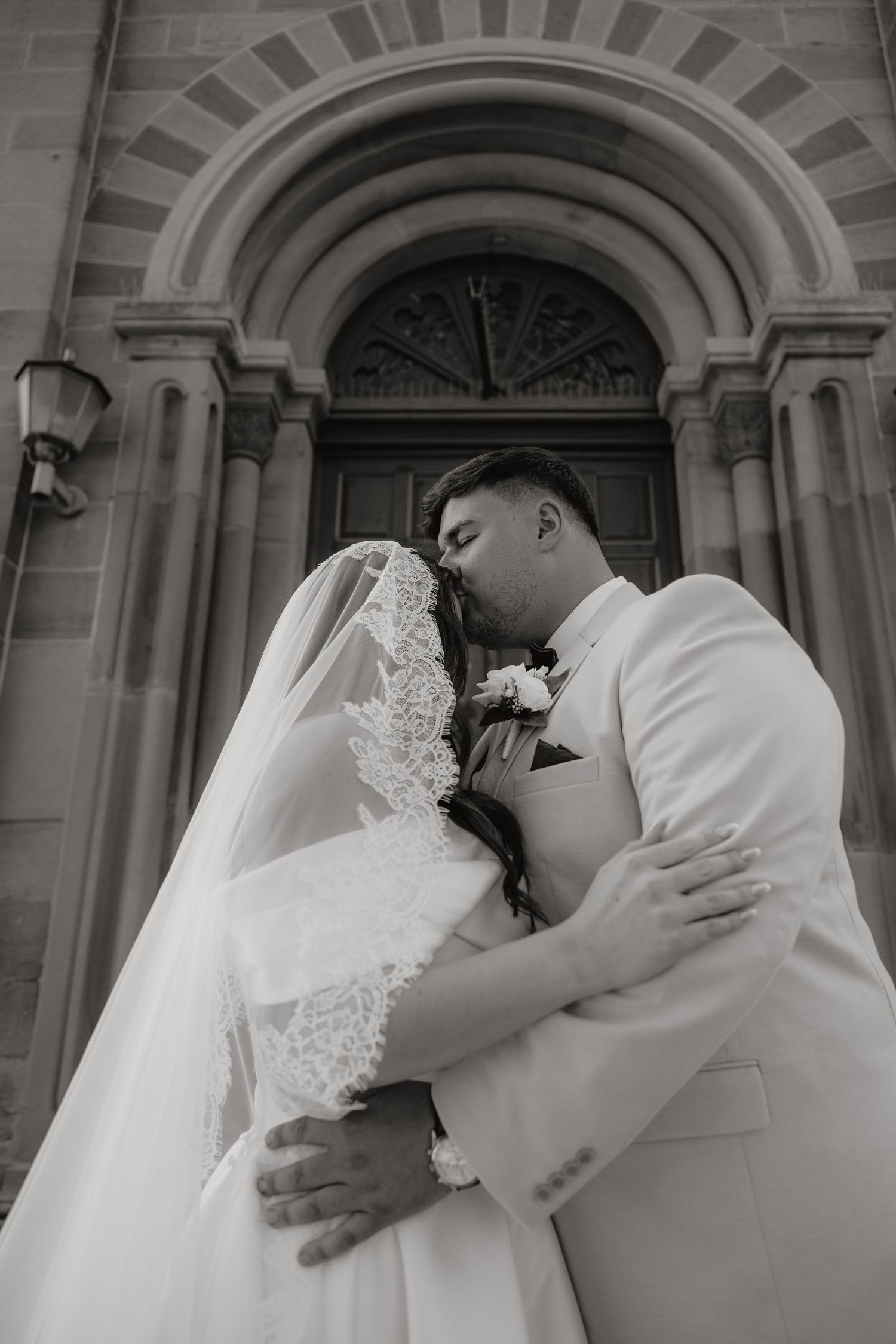 Black and white photo of a bride and groom sharing a romantic kiss outside a historic stone church.