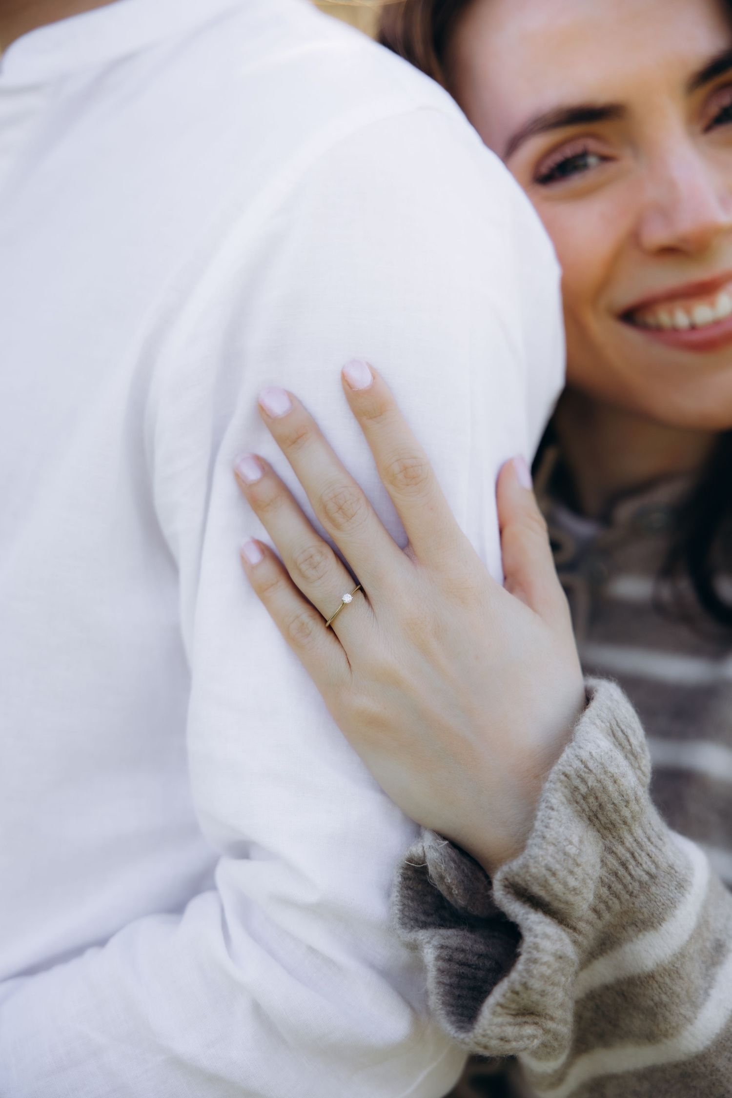 Smiling woman wearing a gold ring hugs a person in a white sweatshirt, showcasing her engagement ring.