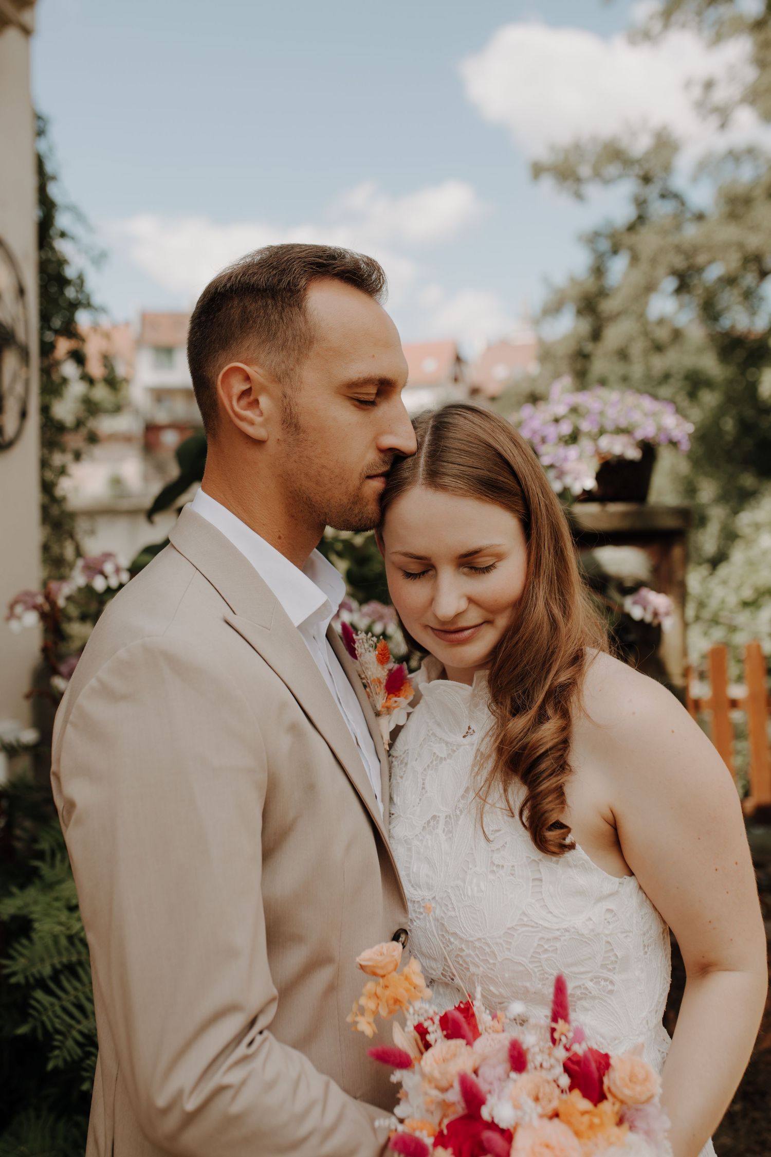 Groom in beige suit gently kisses brides forehead as she holds a vibrant floral bouquet outdoors.
