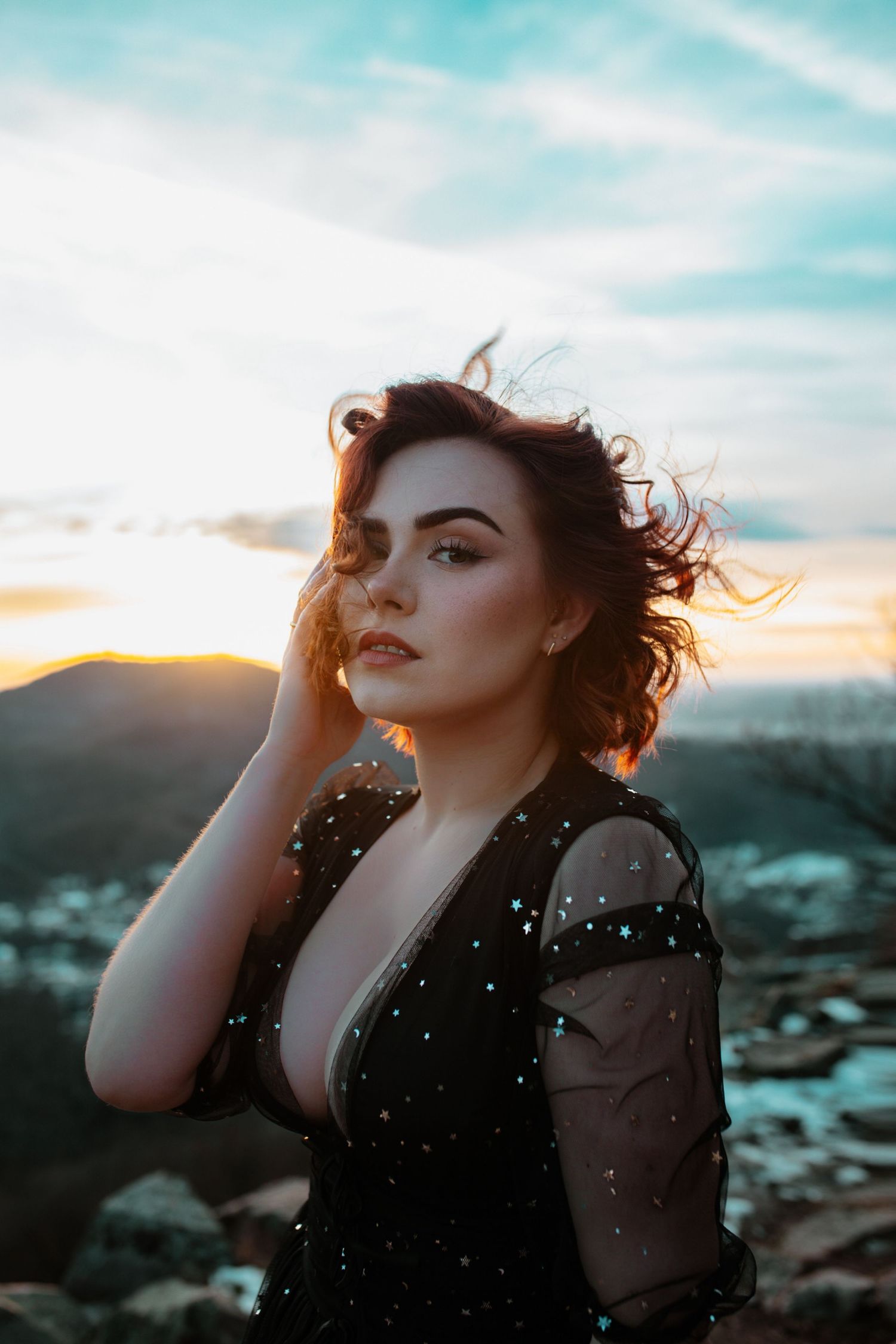 A woman in a black sparkle dress poses at sunset with wind-blown hair on a rocky hilltop.
