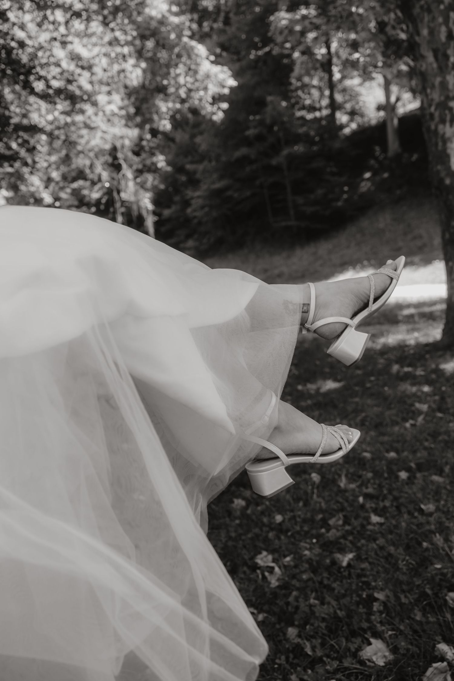 Black and white photo of a bride's legs in elegant strappy heels beneath a flowing wedding dress outdoors.