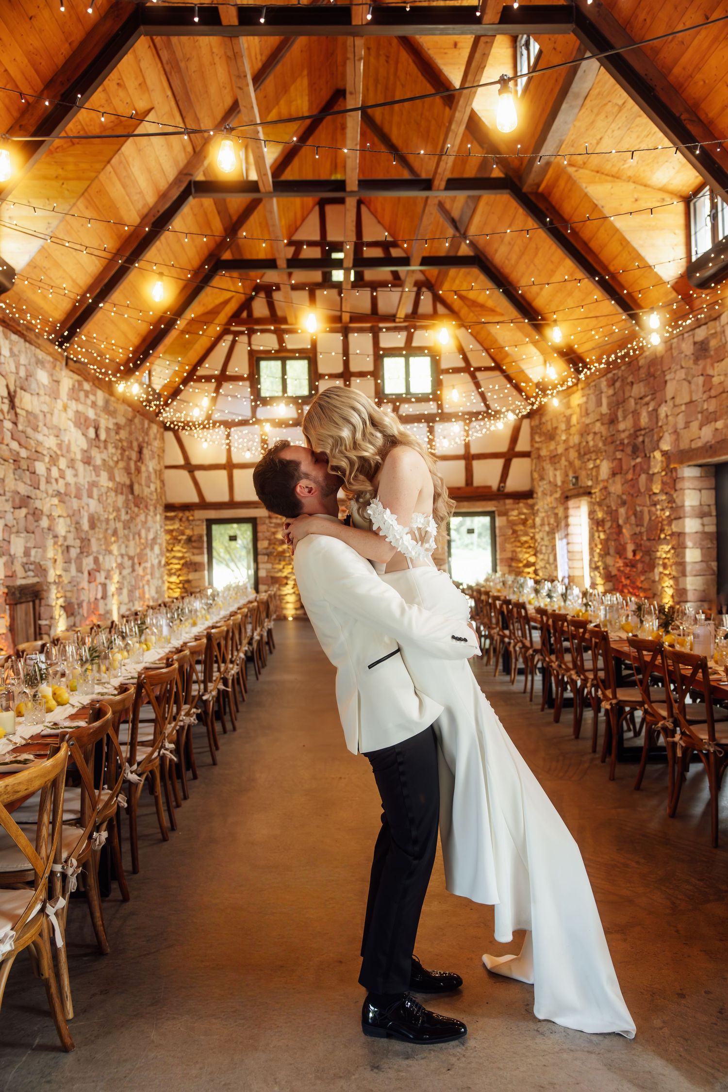 Groom lifts bride in elegant rustic barn wedding venue with string lights, stone walls, and long reception tables.
