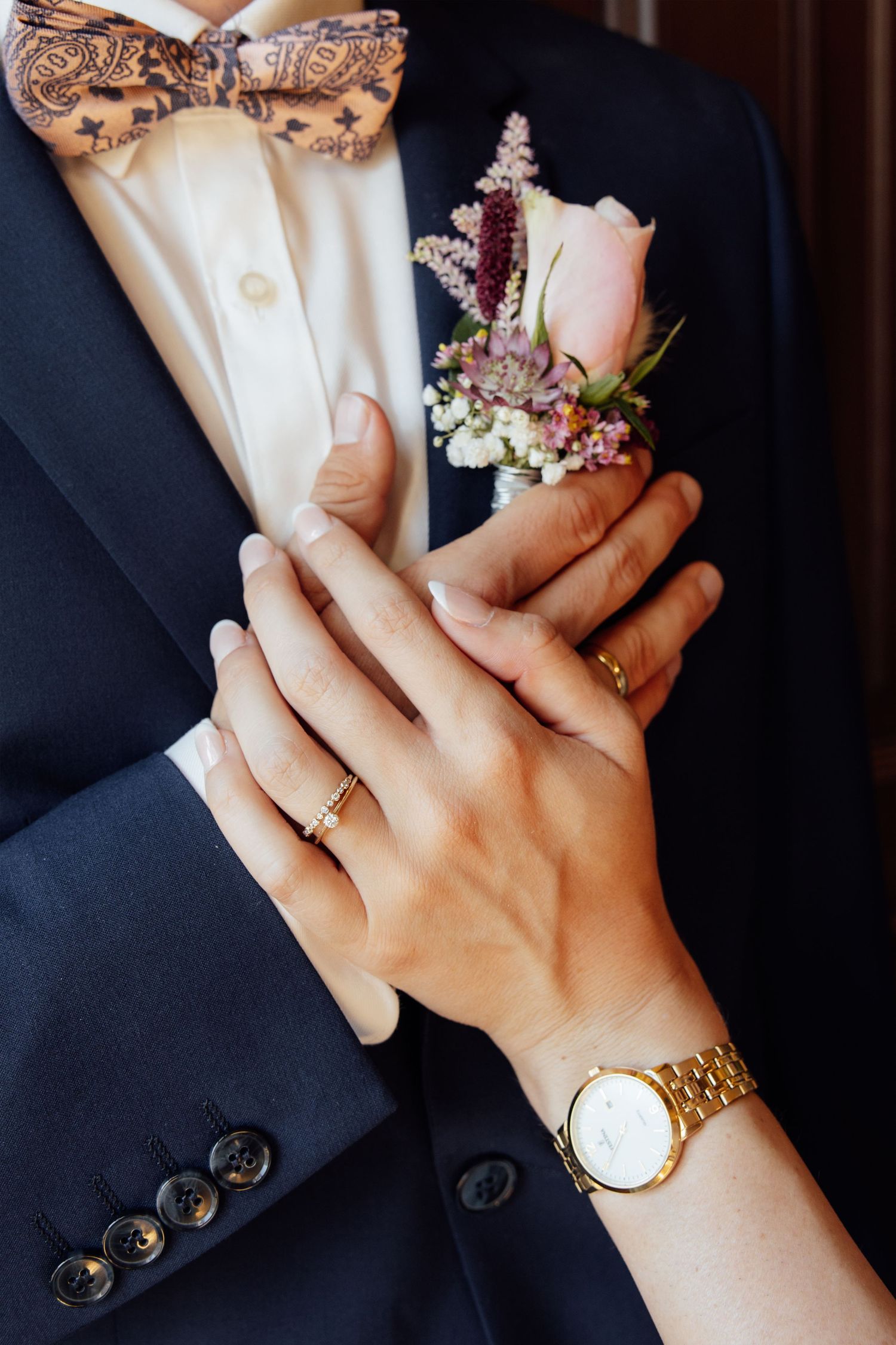 Bride and groom hands with wedding rings, gold watch, boutonniere, navy suit, and patterned bow tie.