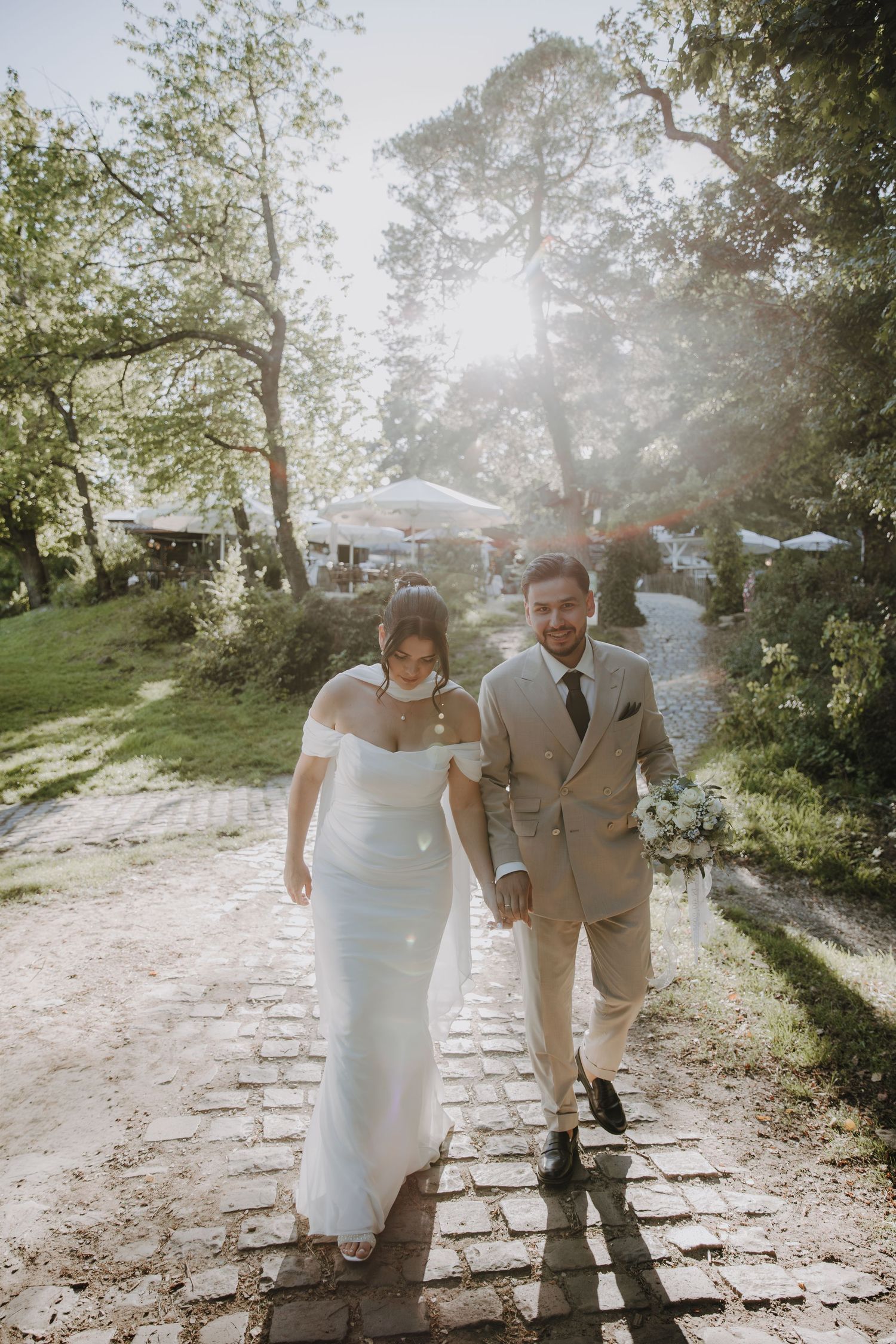 Bride and groom walking hand in hand down sunlit cobblestone path surrounded by trees.