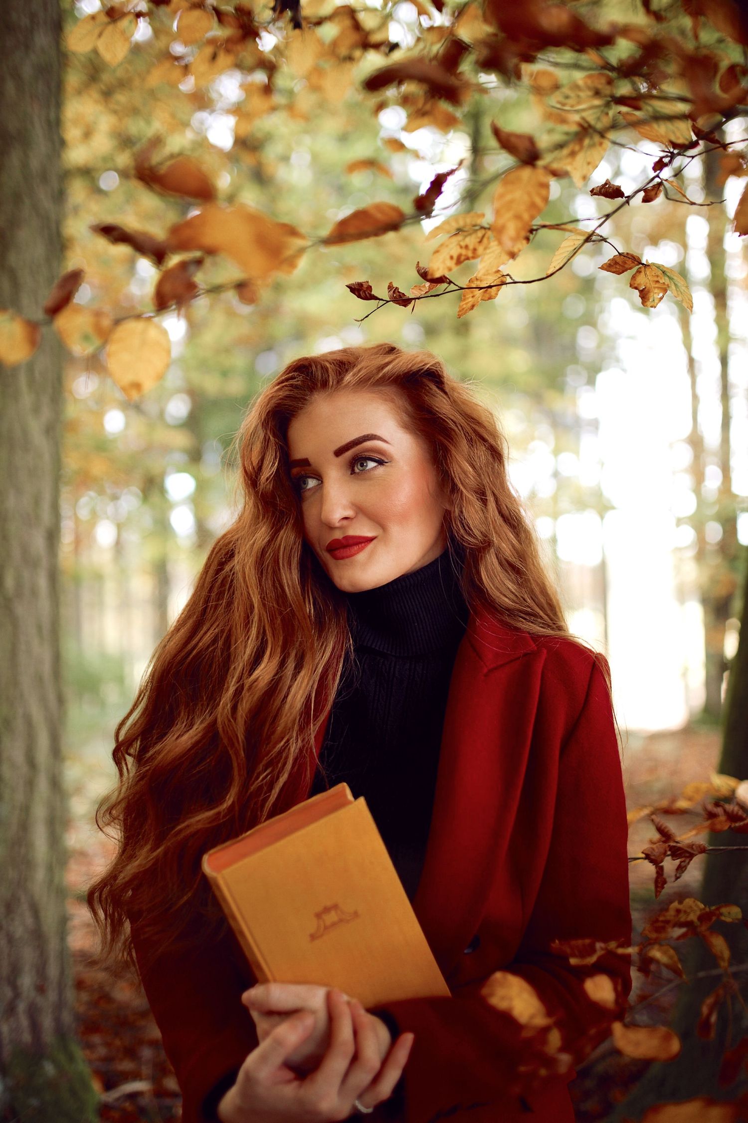 Woman with long red wavy hair holding a book while standing in an autumn forest wearing a red coat.