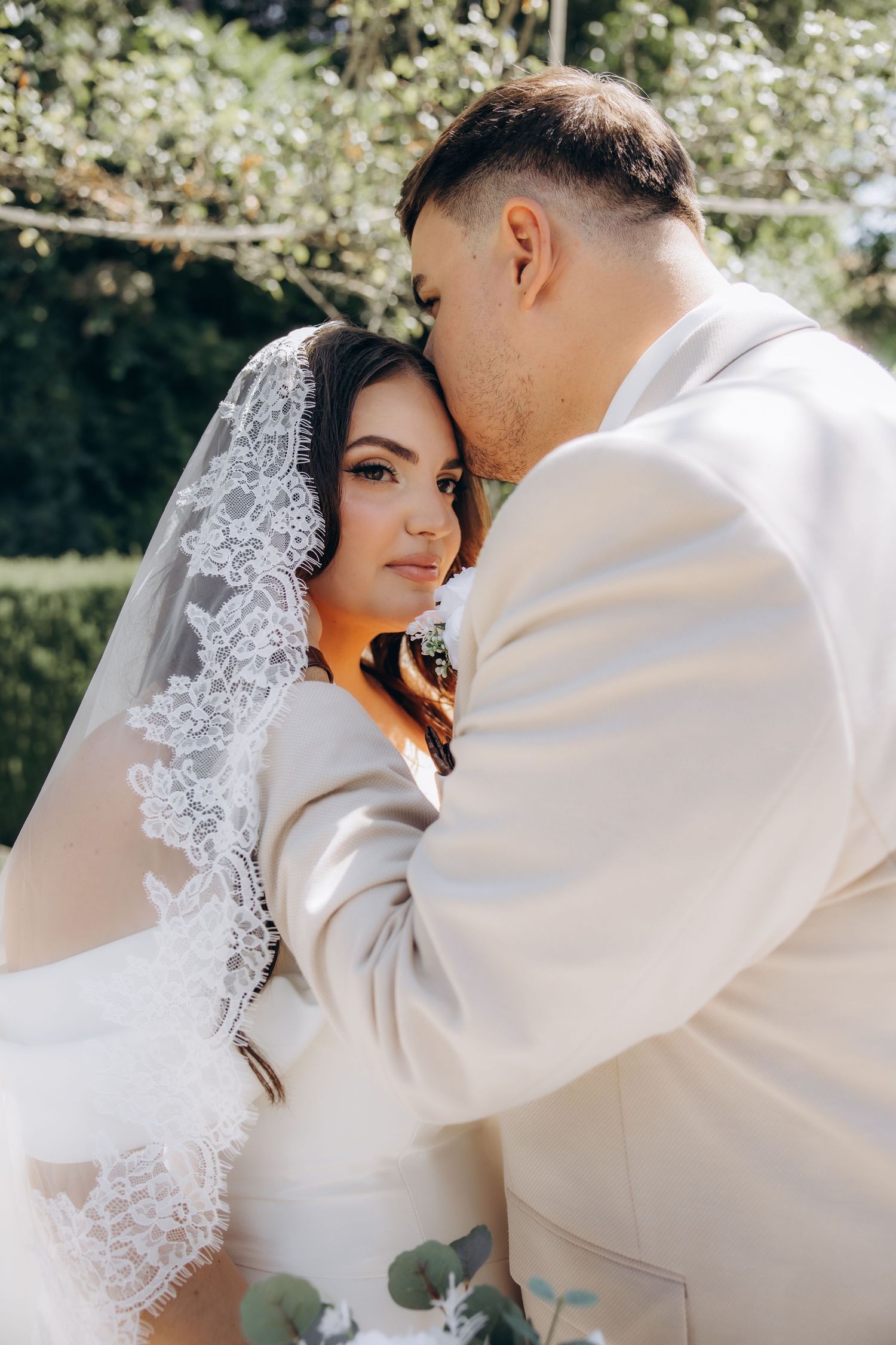Groom in beige suit kissing bride's forehead as she gazes forward in lace veil outdoors.