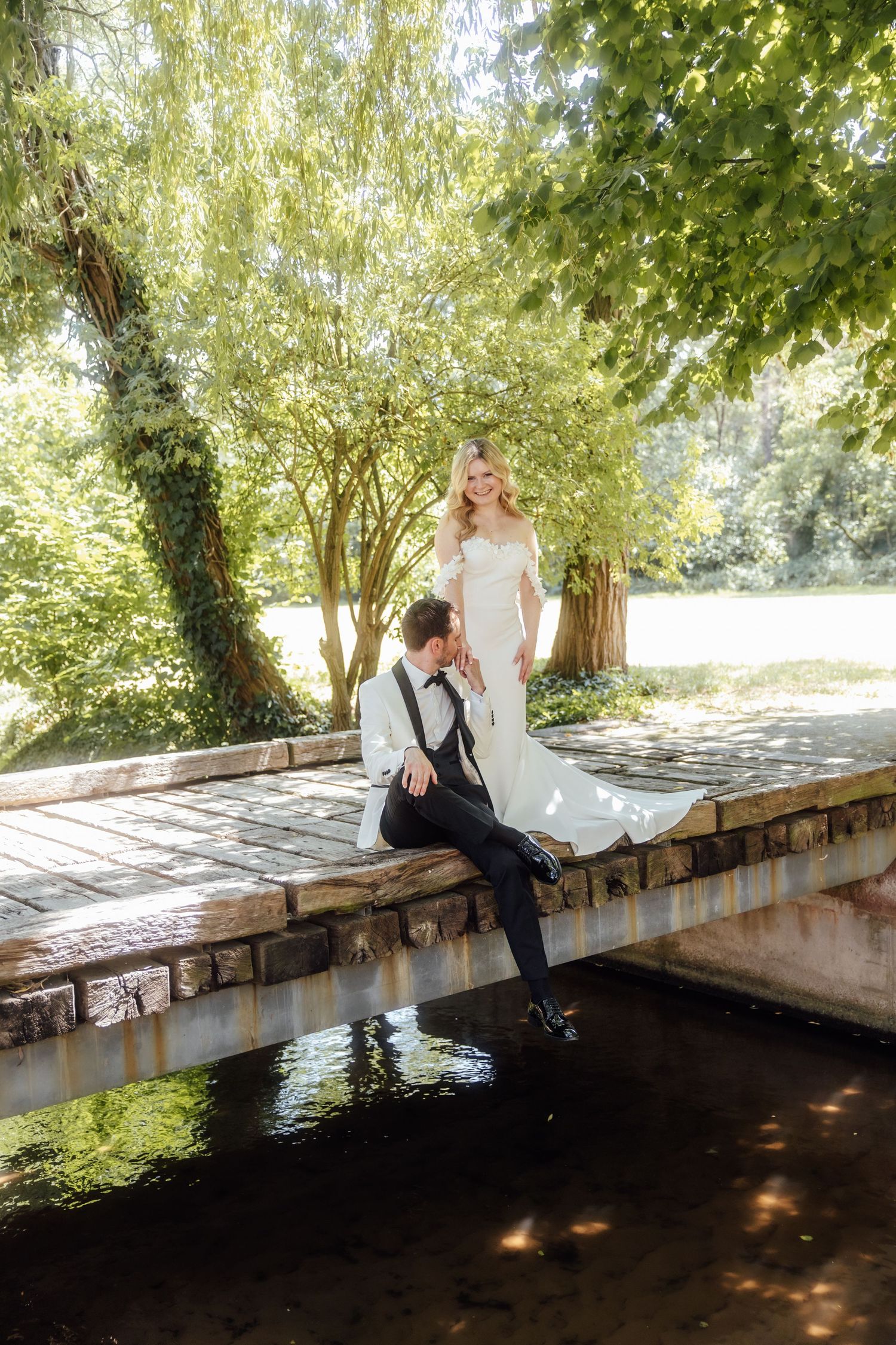 Bride and groom pose on a stone bridge surrounded by lush green trees on their wedding day.