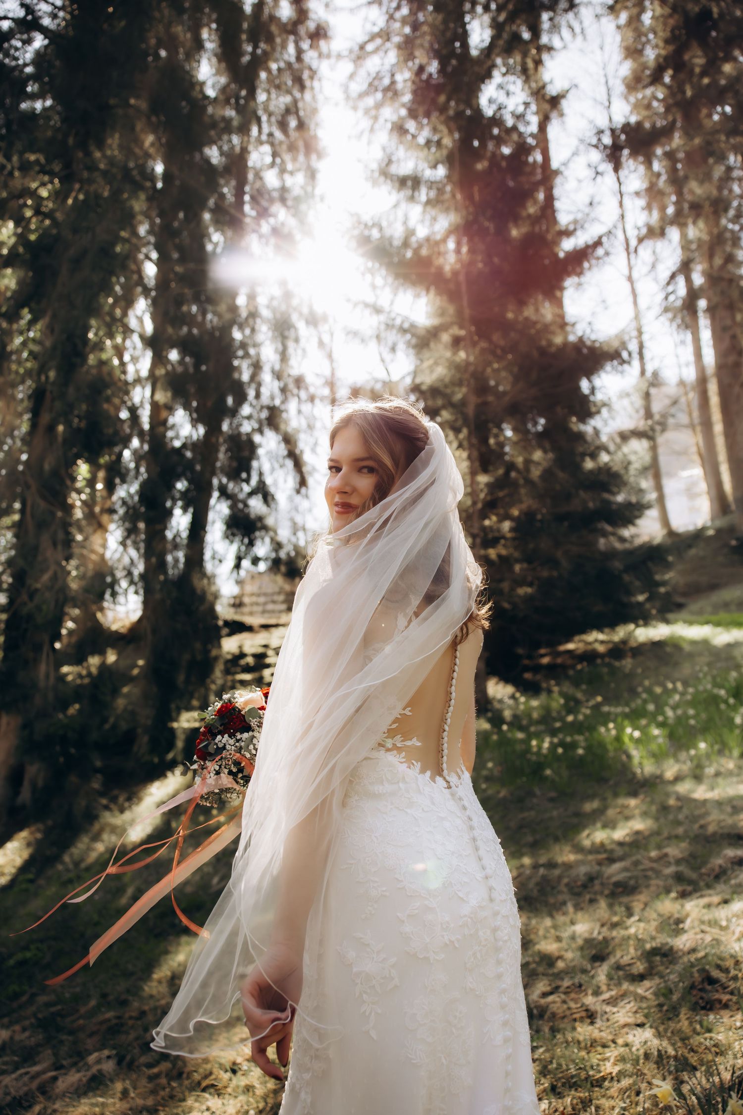 Bride in lace wedding dress with veil looking back over shoulder in sunlit forest.