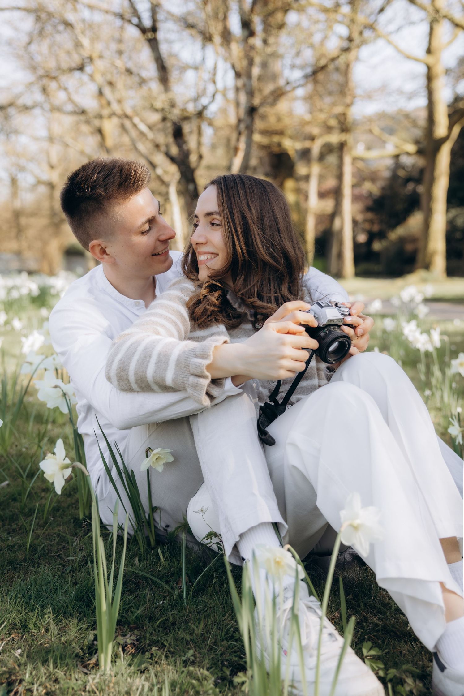 A couple sits together in a spring park, smiling and holding a camera among white daffodils, both dressed in white.