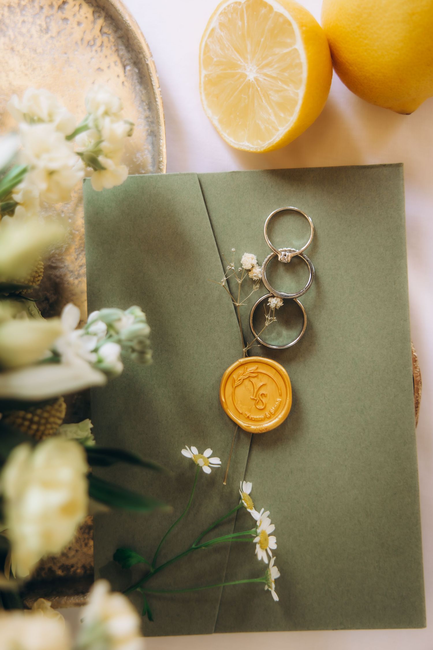 Green envelope with gold wax seal, wedding rings, daisies, and lemons on white surface.