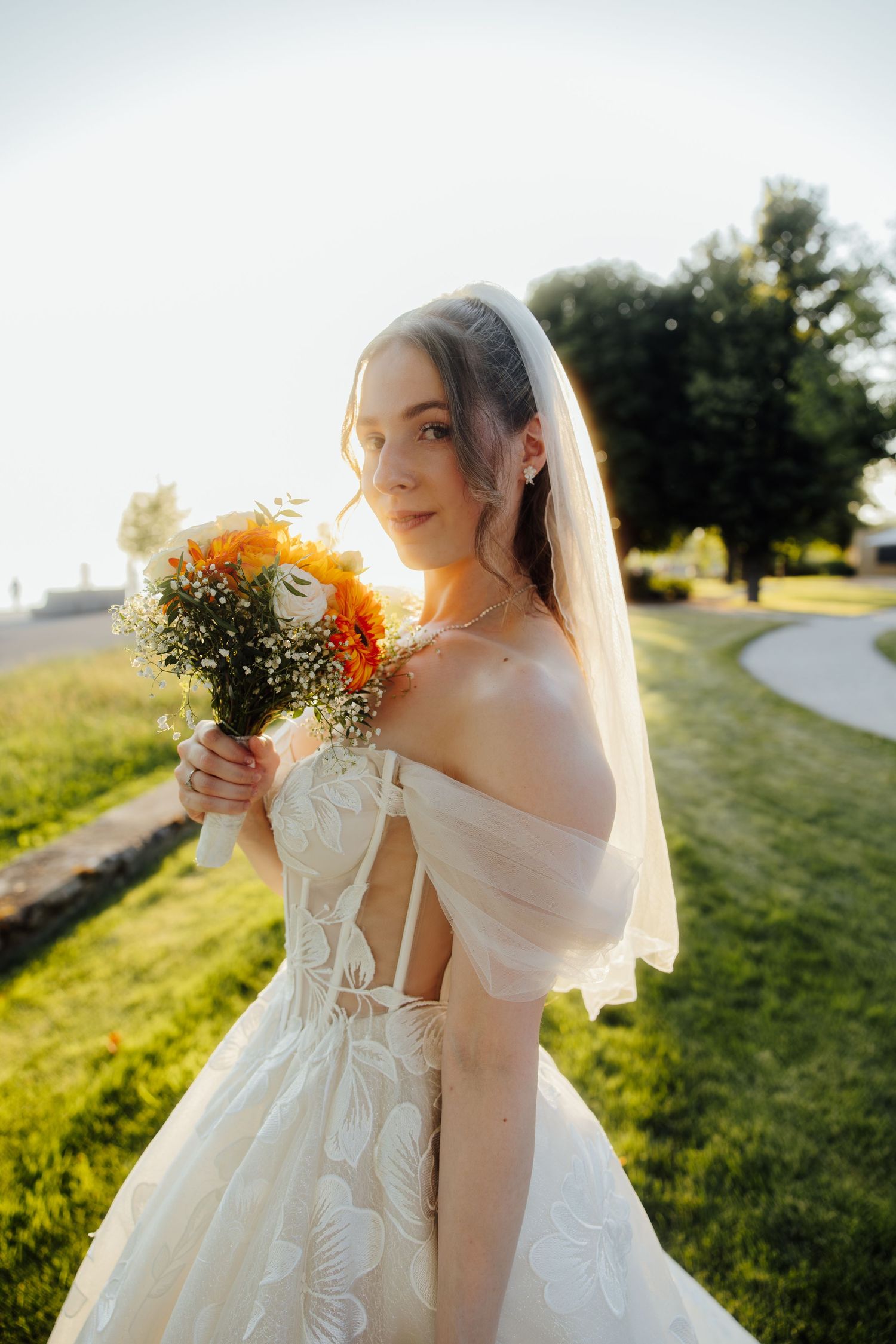 Bride in floral lace gown holding colorful bouquet outdoors in golden sunlight.