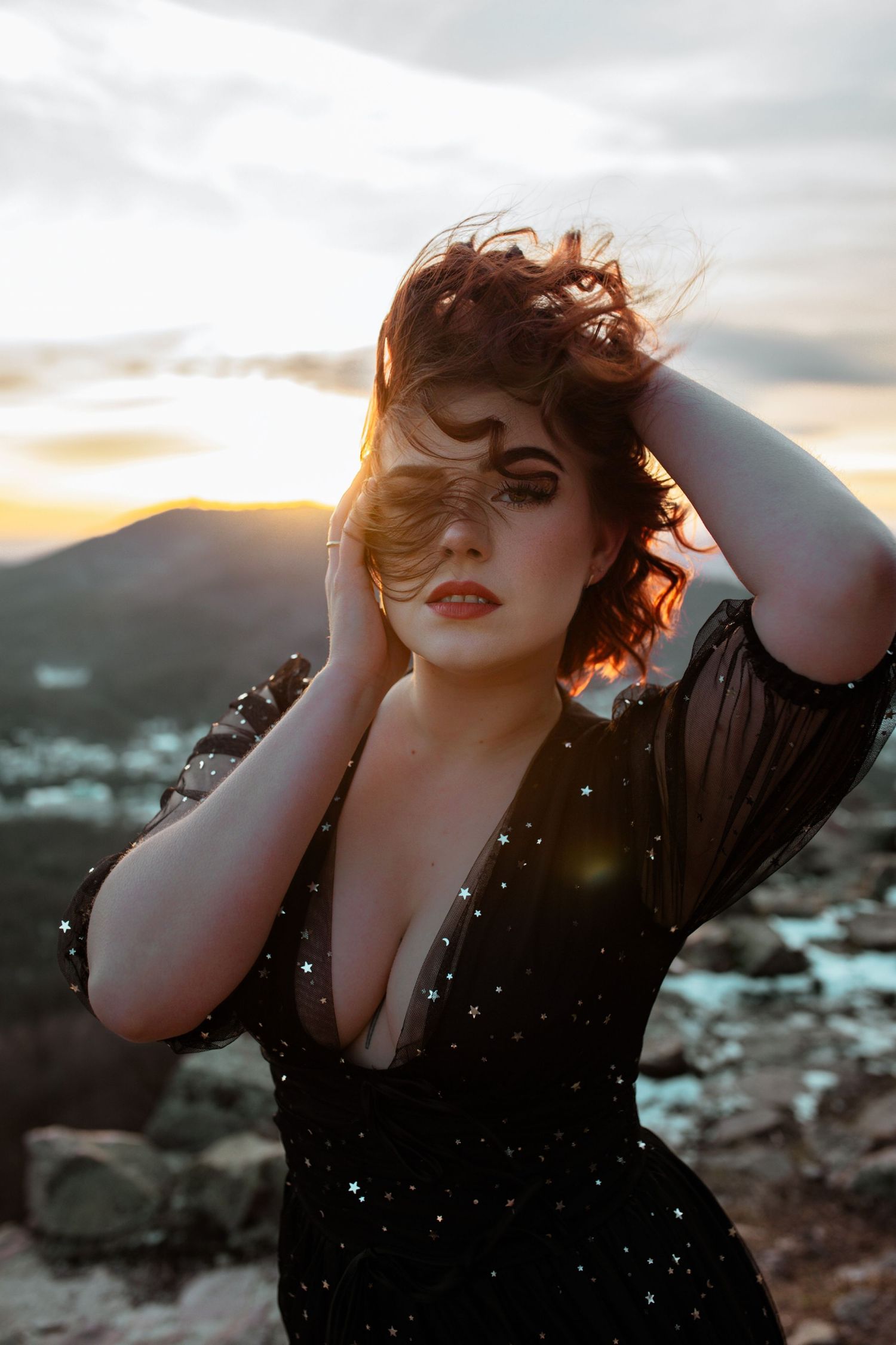 A curvy woman in a black sequined dress poses dramatically at sunset on a rocky mountain.