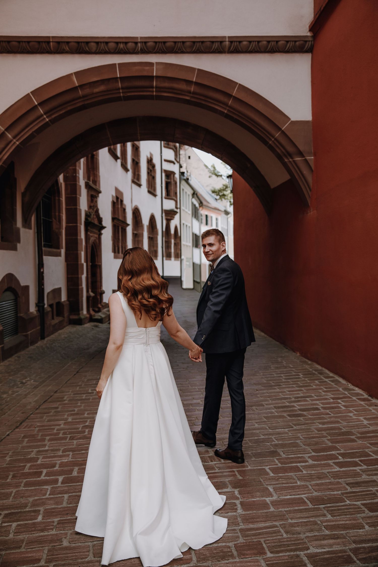 Bride and groom walk hand in hand through a charming cobblestone archway in a historic town.