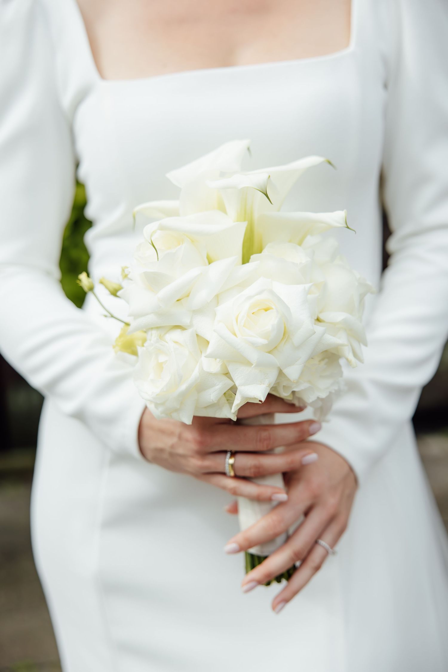 Bride in white long-sleeve gown holding elegant bouquet of white roses and calla lilies, wearing gold wedding band.