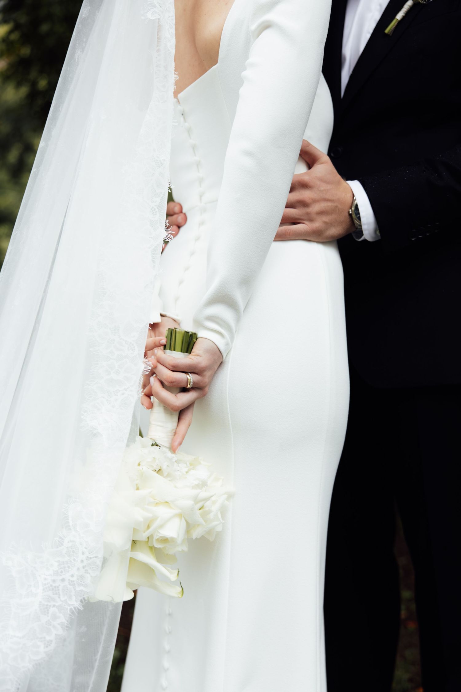 Bride in white gown holding bouquet of white roses, embraced by groom in black suit at wedding ceremony.