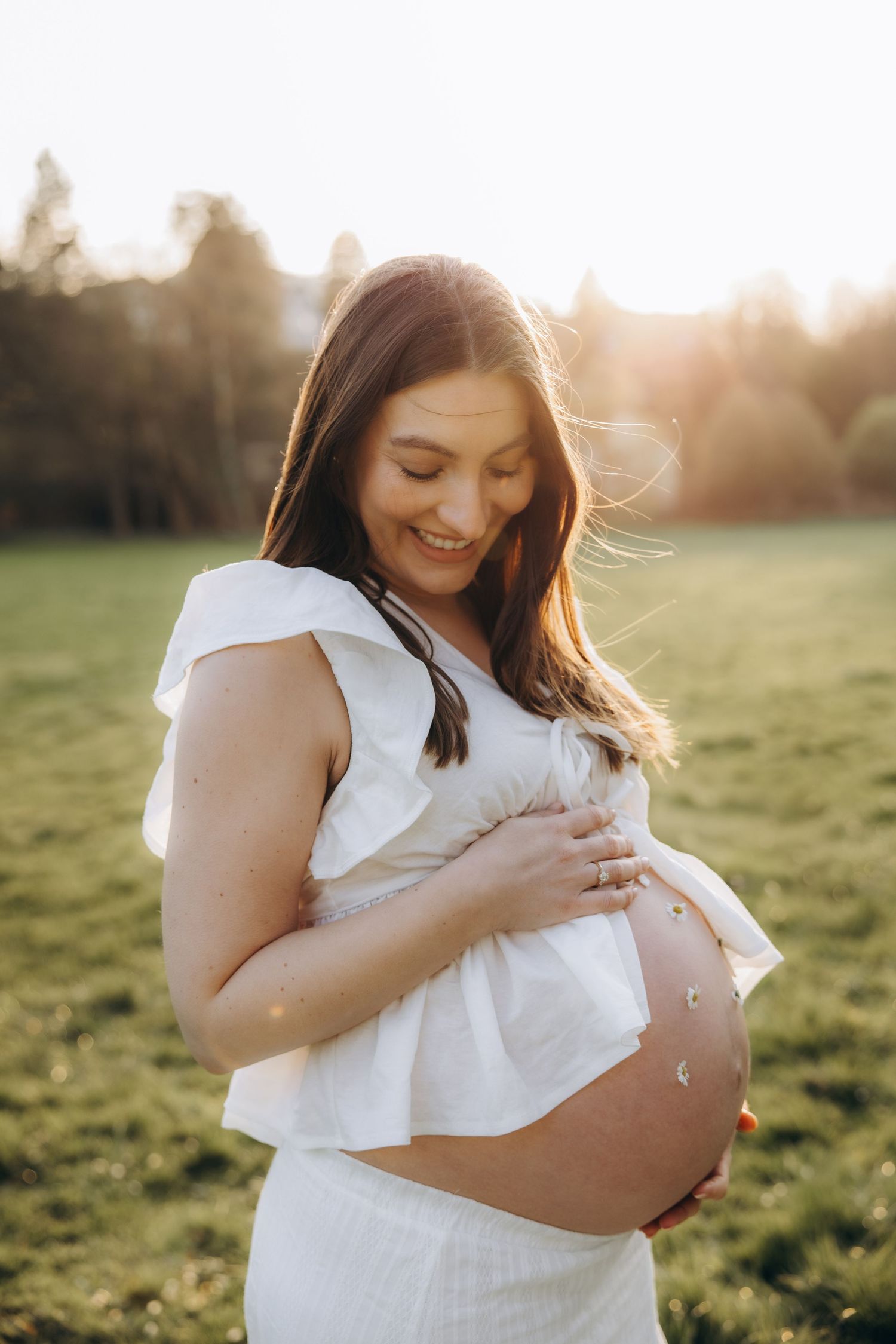 Smiling pregnant woman in white dress holding baby bump in golden sunset field.