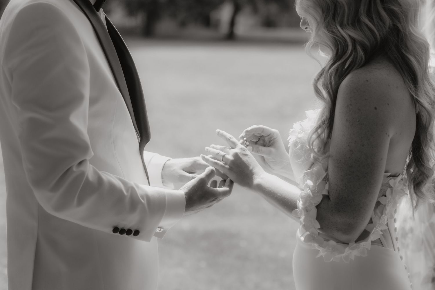 Black and white photo of a couple exchanging wedding rings during an outdoor ceremony.
