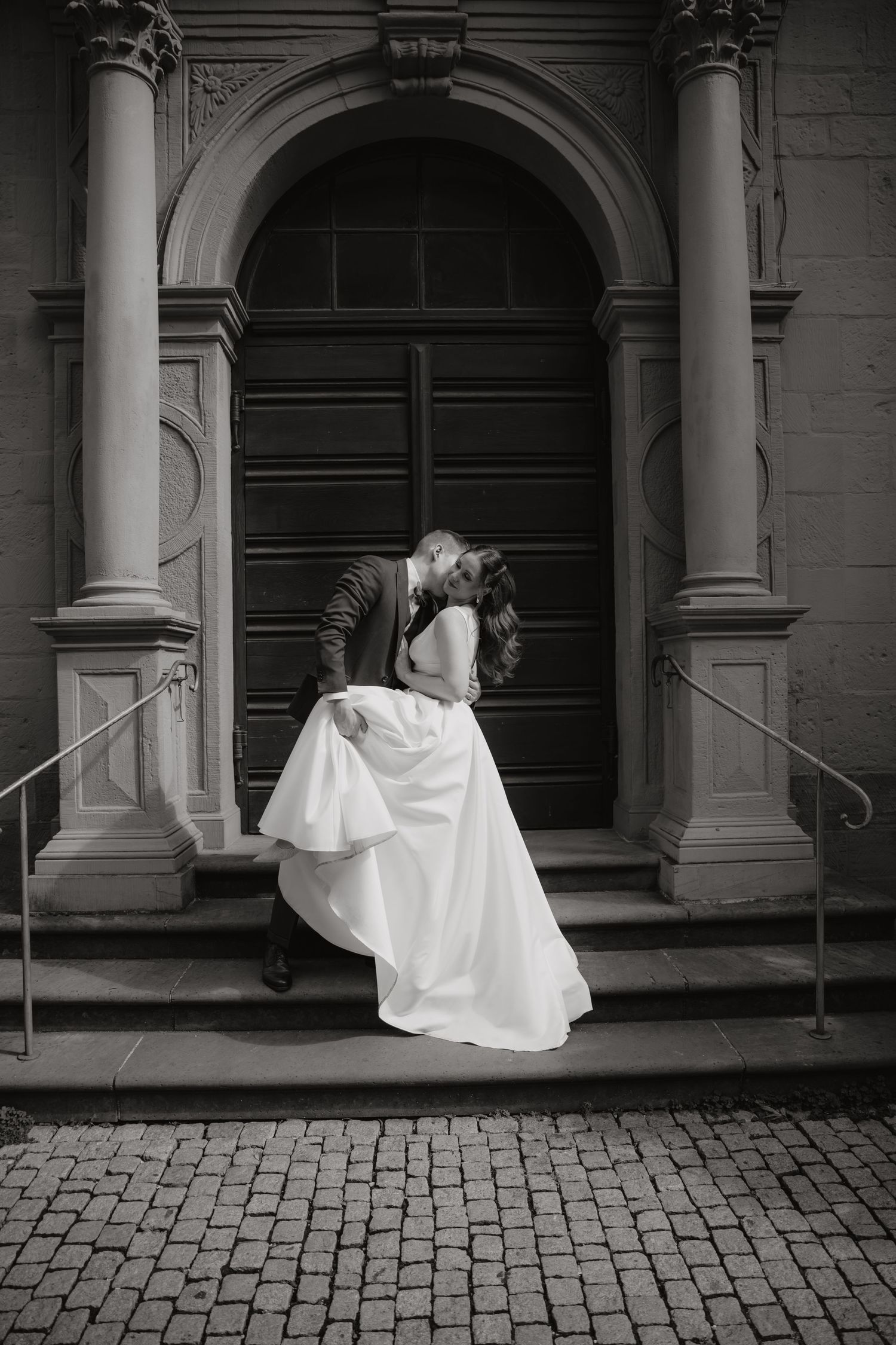 Bride and groom share a romantic dip kiss on steps of an ornate church doorway in black and white.