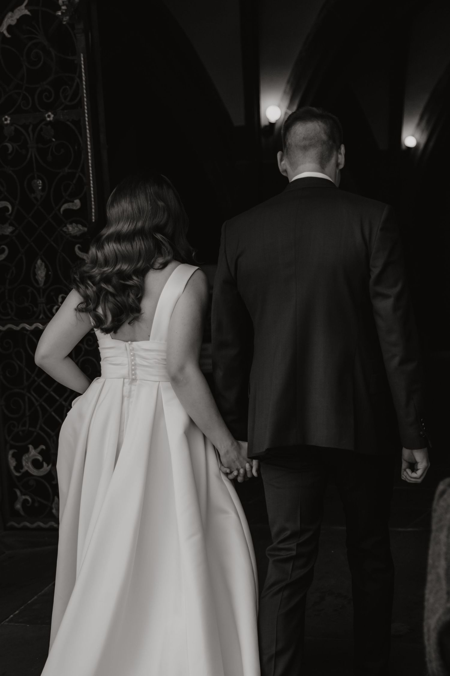 Black and white photo of bride and groom holding hands walking toward ornate dark doorway.