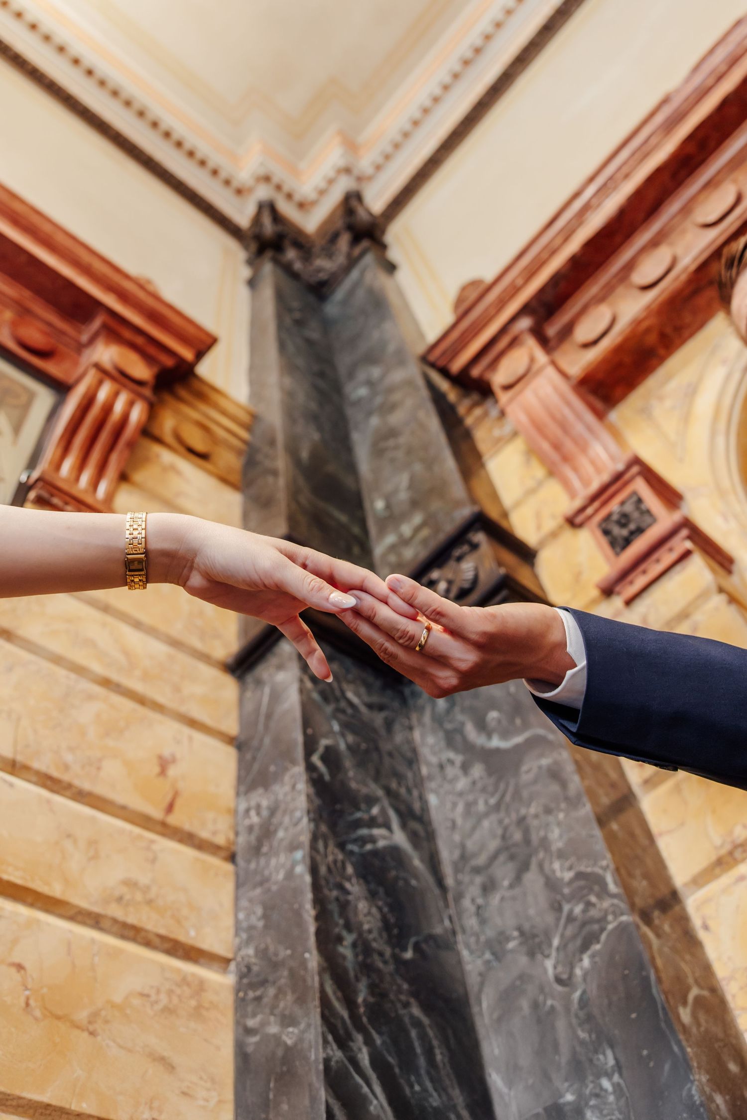 Couple exchanging wedding rings in an elegant marble columned hall, close-up of hands with gold bracelet and ring.