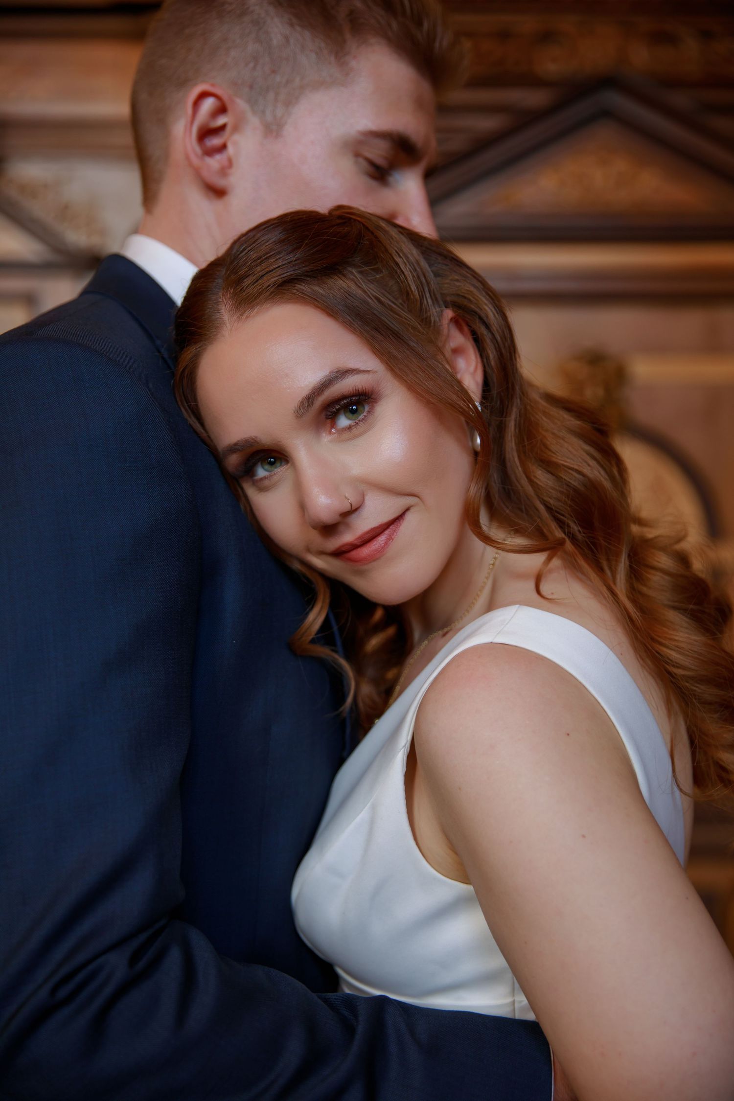 Smiling bride with wavy auburn hair leans against groom in navy suit near carved wooden door.