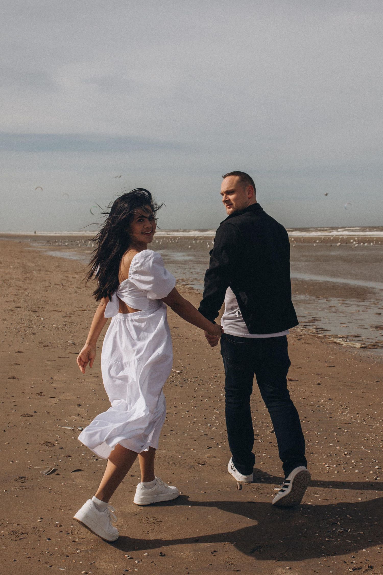 Couple walks hand in hand along a windy beach, bride wearing a flowing white dress.