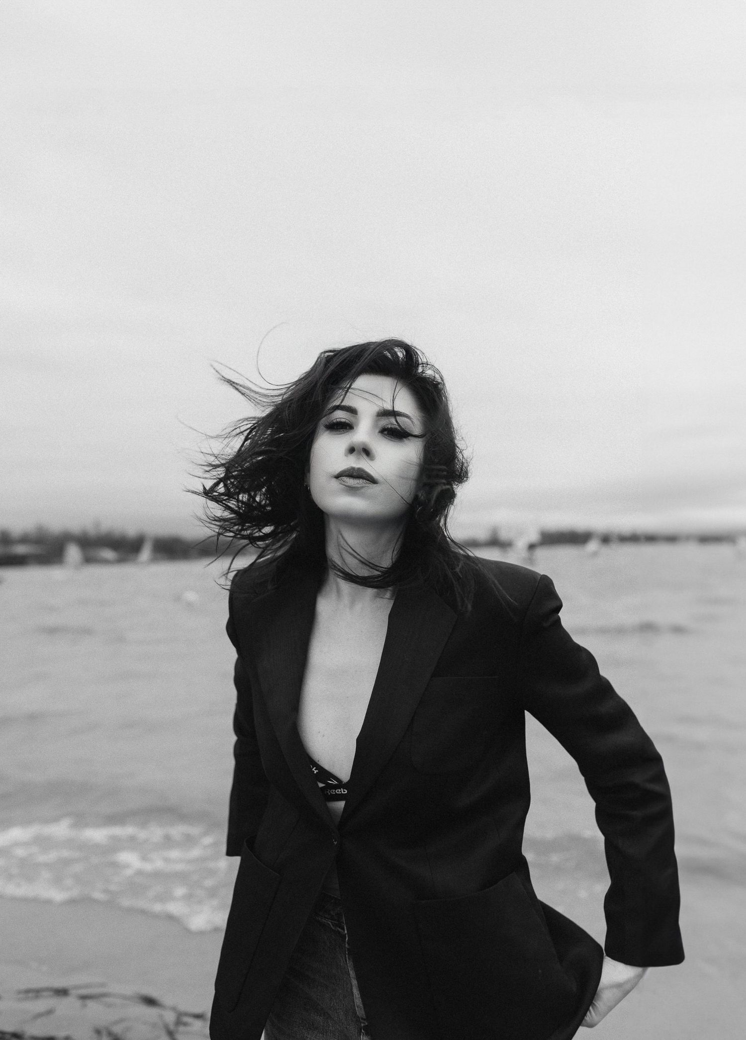 Black and white portrait of a dark-haired woman in a blazer posing on a windy seaside beach.