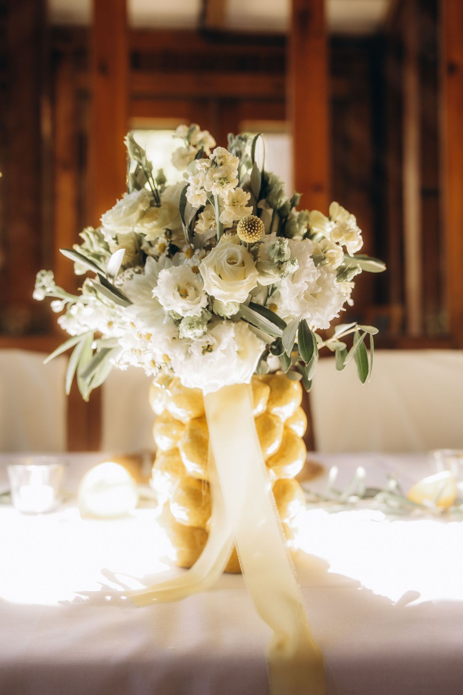 Elegant white floral wedding centerpiece with cream ribbon on a white tablecloth in a rustic venue.