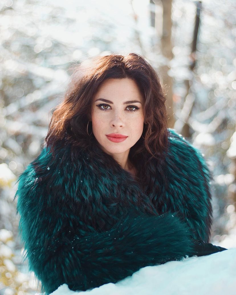 A brunette woman in a teal fur coat poses in a bright snowy outdoor setting.