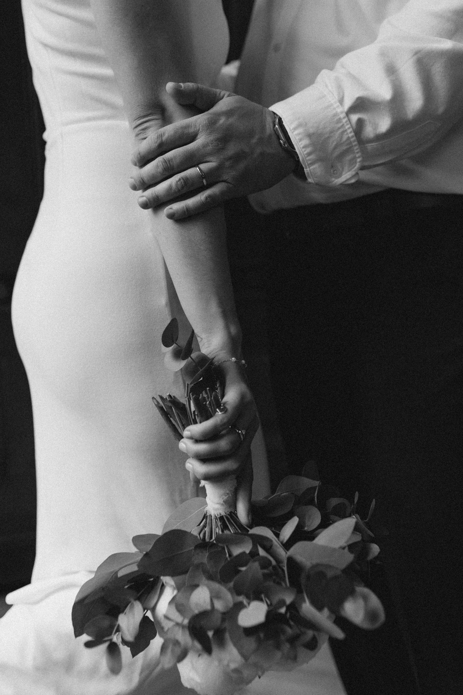 Black and white photo of groom embracing bride from behind while she holds eucalyptus wedding bouquet.