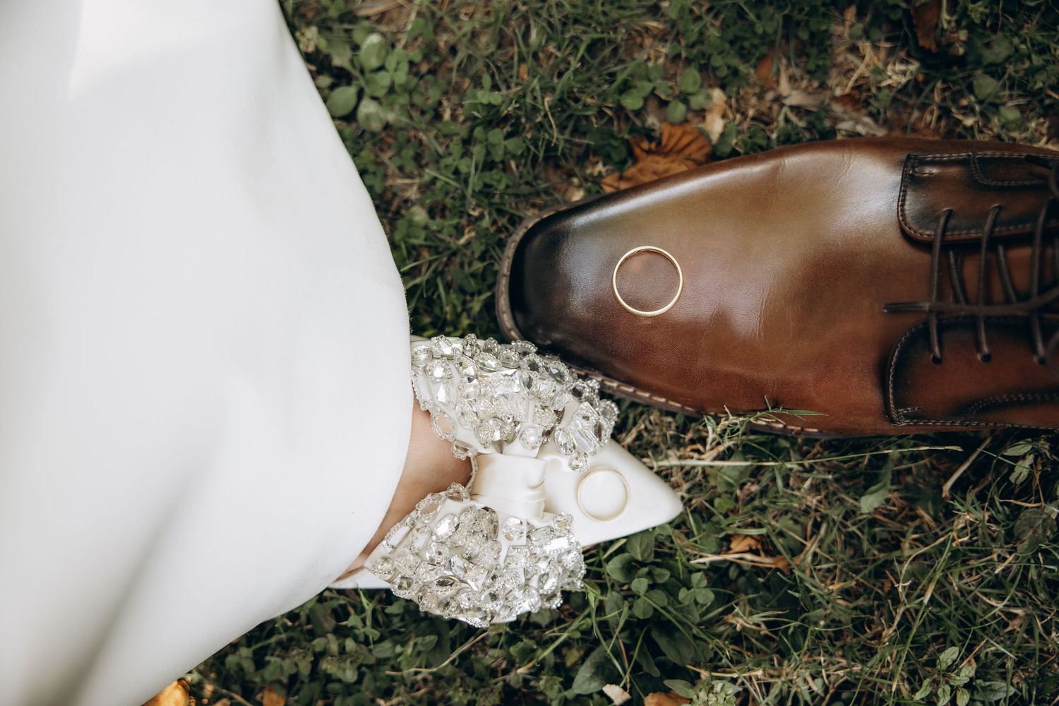 Wedding rings resting on a brown leather dress shoe beside a white lace bridal gown on green grass.
