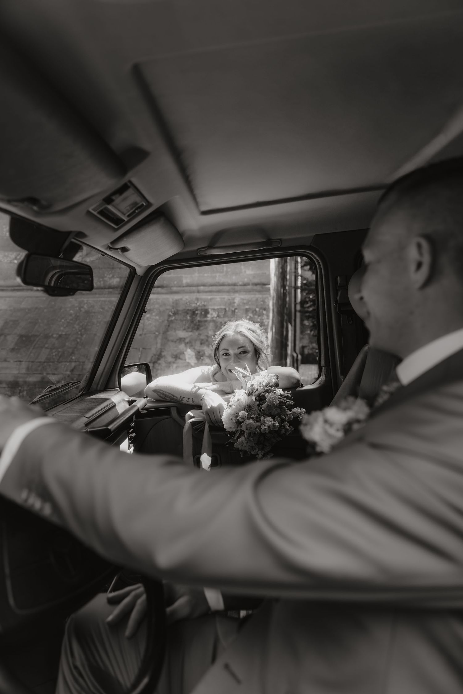 Black and white photo of a bride leaning into a vintage car window, smiling at a suited man holding a bouquet.